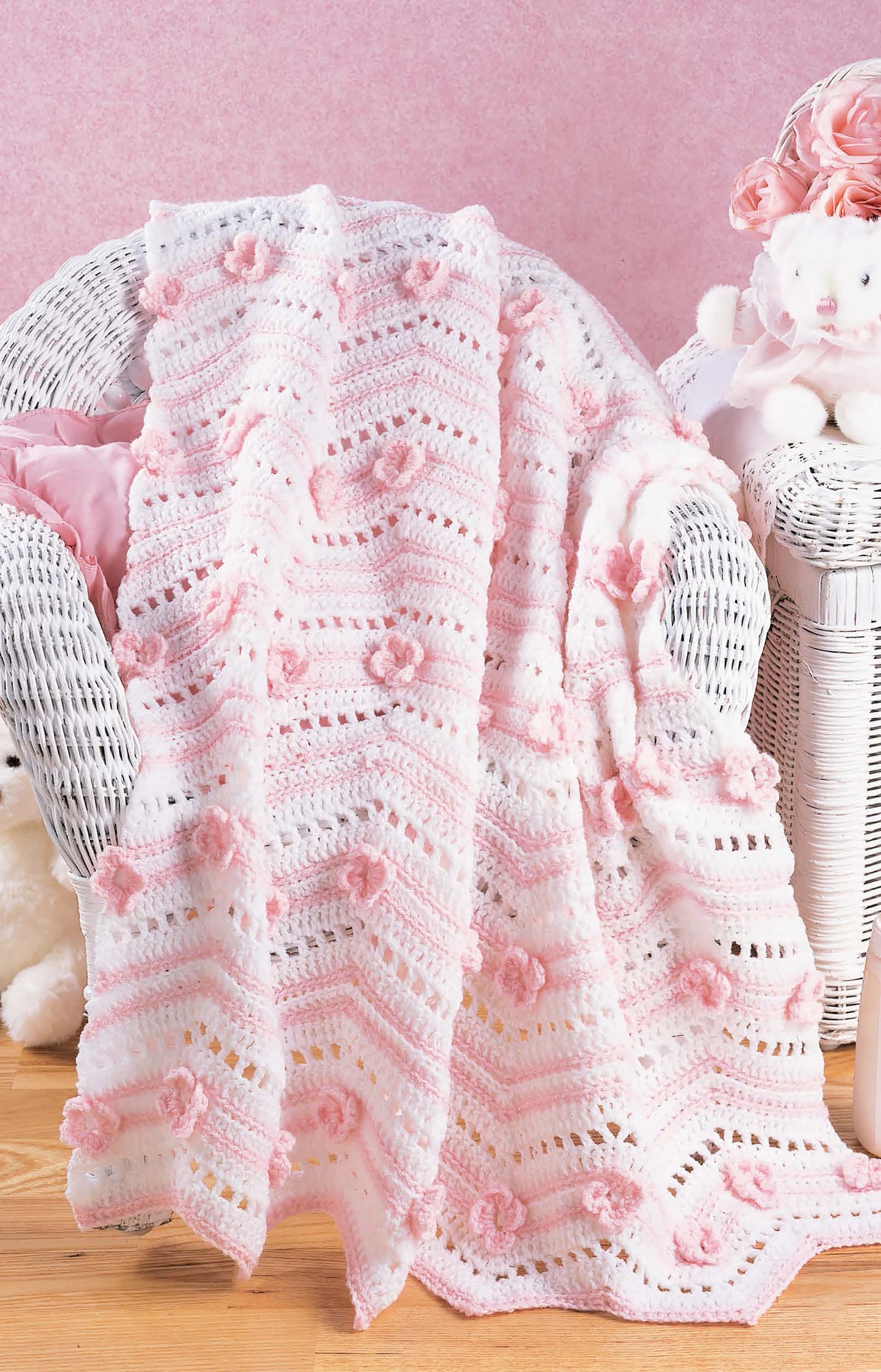A cozy, feminine room features a white and pink crocheted blanket made with the Learn to Crochet Ripple Afghans Digital Download from Leisure Arts, draped on a wicker chair beside a plush teddy bear and pastel pink decor.