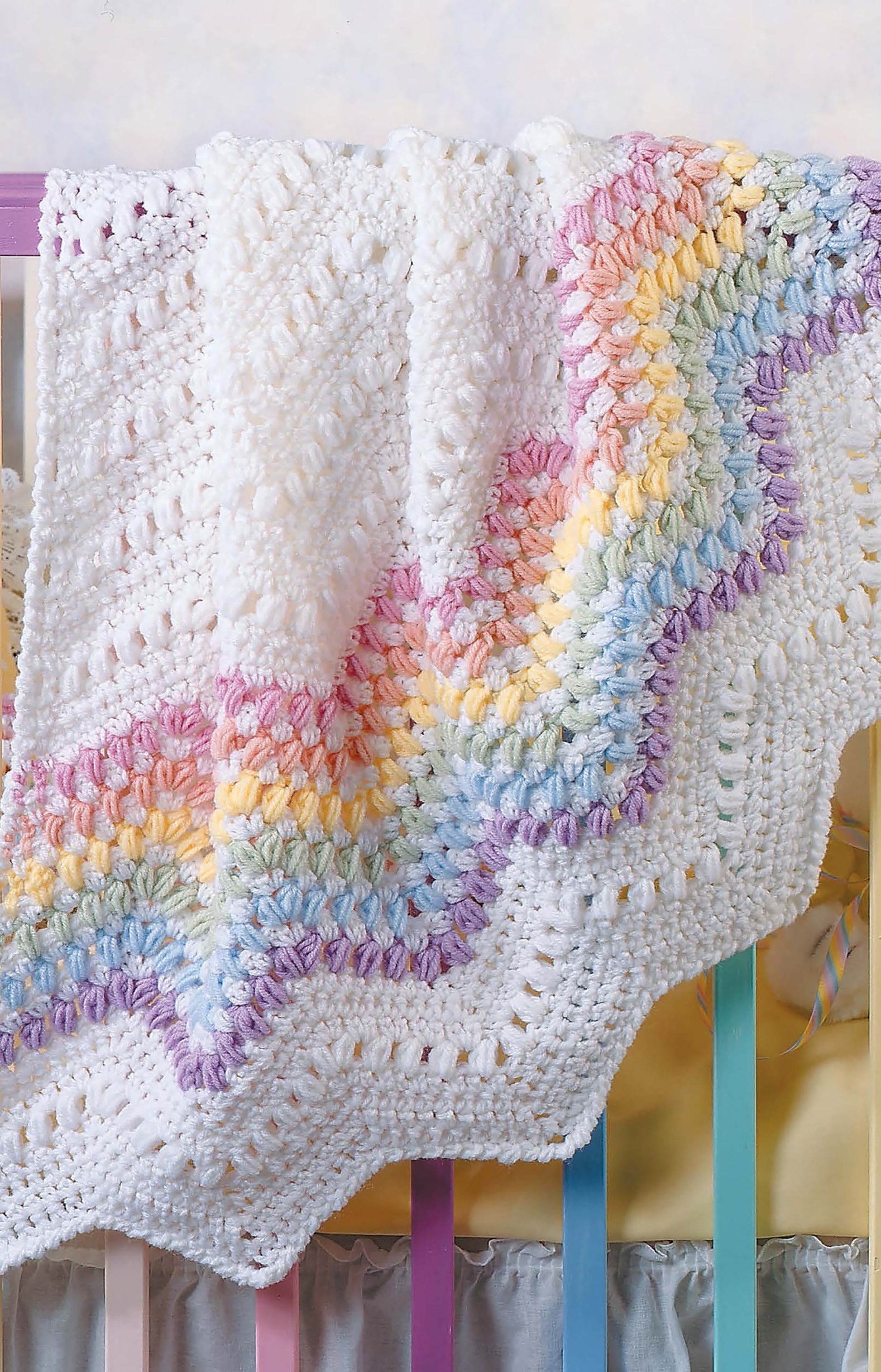 A crocheted blanket made with Leisure Arts' Learn to Crochet Ripple Afghans Digital Download features soft pastel rainbow and white zigzag patterns, draped over a crib with pastel-painted bars that complement its cozy, colorful look.