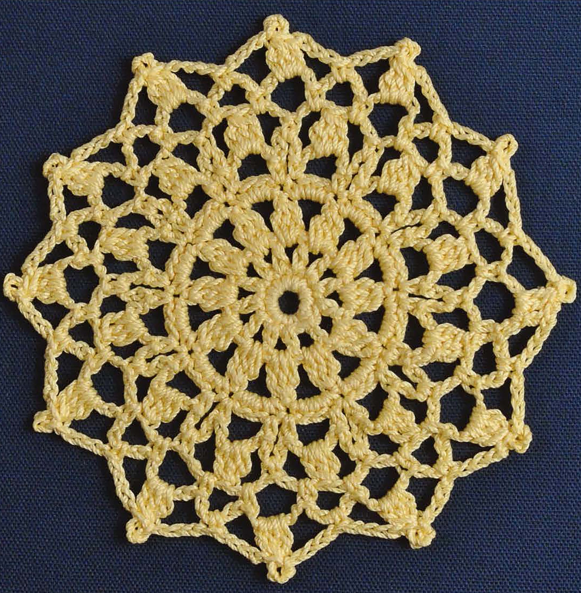 Showcased on a dark blue background, this yellow doily with a flower-like pattern and twelve points demonstrates advanced crochet techniques—great inspiration from Leisure Arts’ “Complete Guide to Symbol Crochet.”.