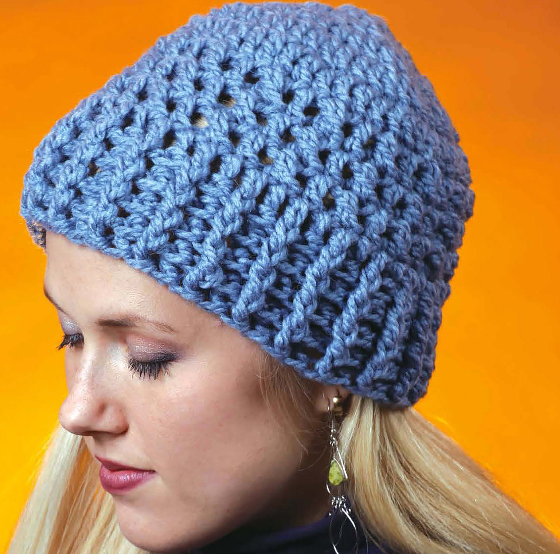 A woman with long blonde hair wears a chunky, light blue crocheted beanie—made with techniques from the Complete Guide to Symbol Crochet by Leisure Arts—and silver earrings, posing against an orange background with her eyes closed.