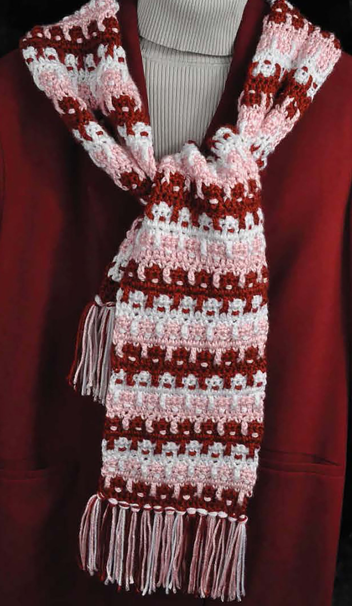 The Complete Guide to Symbol Crochet by Leisure Arts is displayed on a mannequin draped in a red and pink crocheted scarf with white accents and fringe, paired with a maroon coat and light ribbed turtleneck sweater.