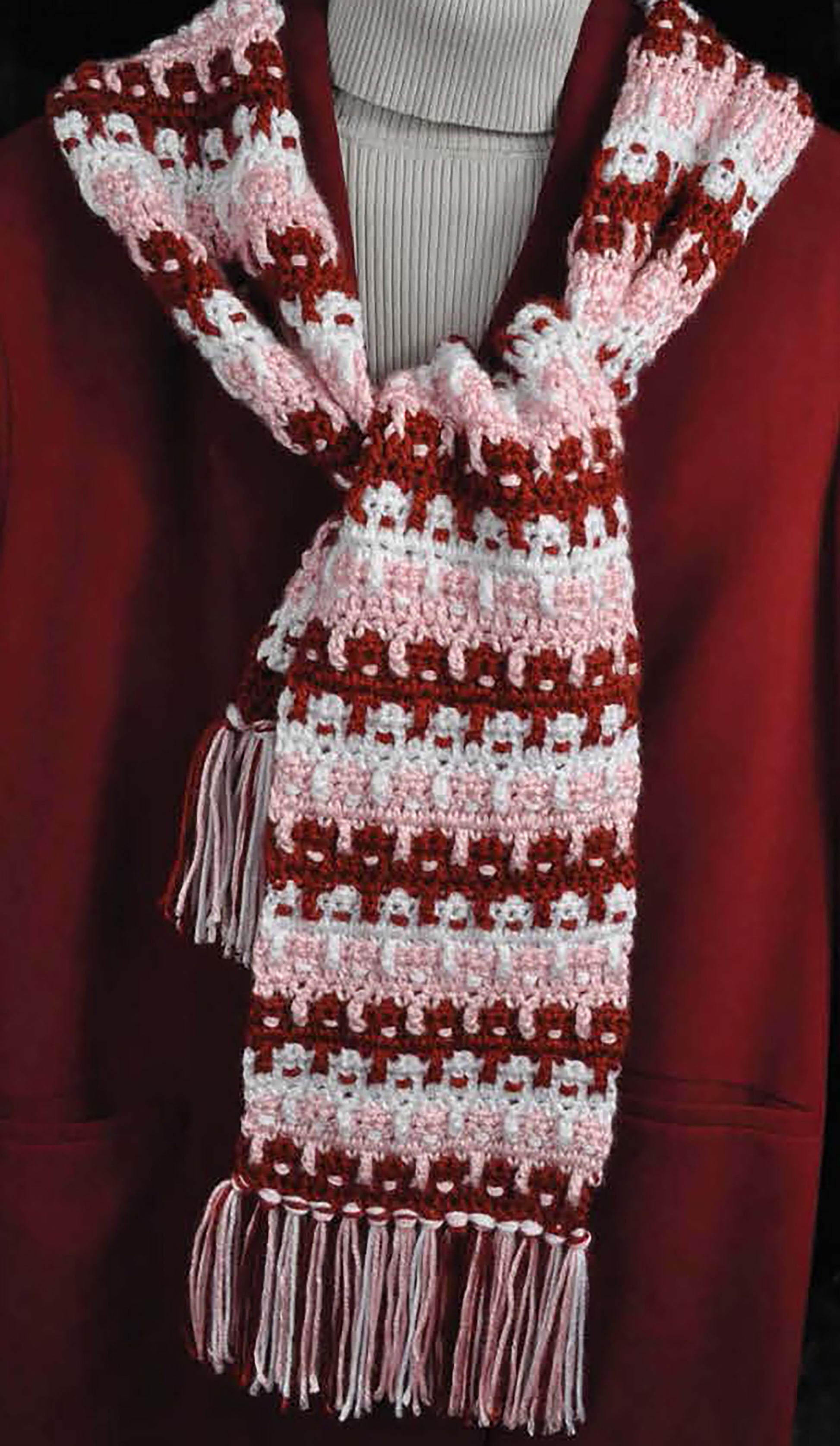 The Complete Guide to Symbol Crochet by Leisure Arts is displayed on a mannequin draped in a red and pink crocheted scarf with white accents and fringe, paired with a maroon coat and light ribbed turtleneck sweater.