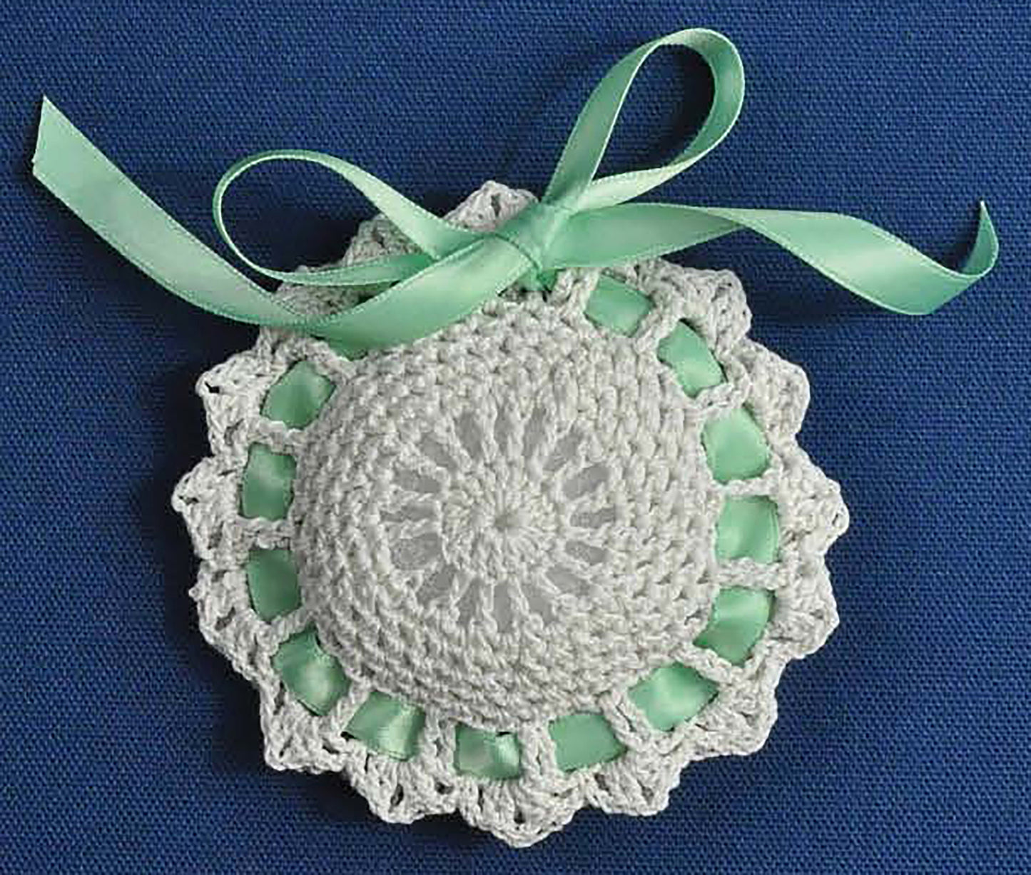 Displayed on a blue fabric background, the round white crocheted ornament with scalloped edges is perfect inspiration for beginner projects featured in the Complete Guide to Symbol Crochet by Leisure Arts.