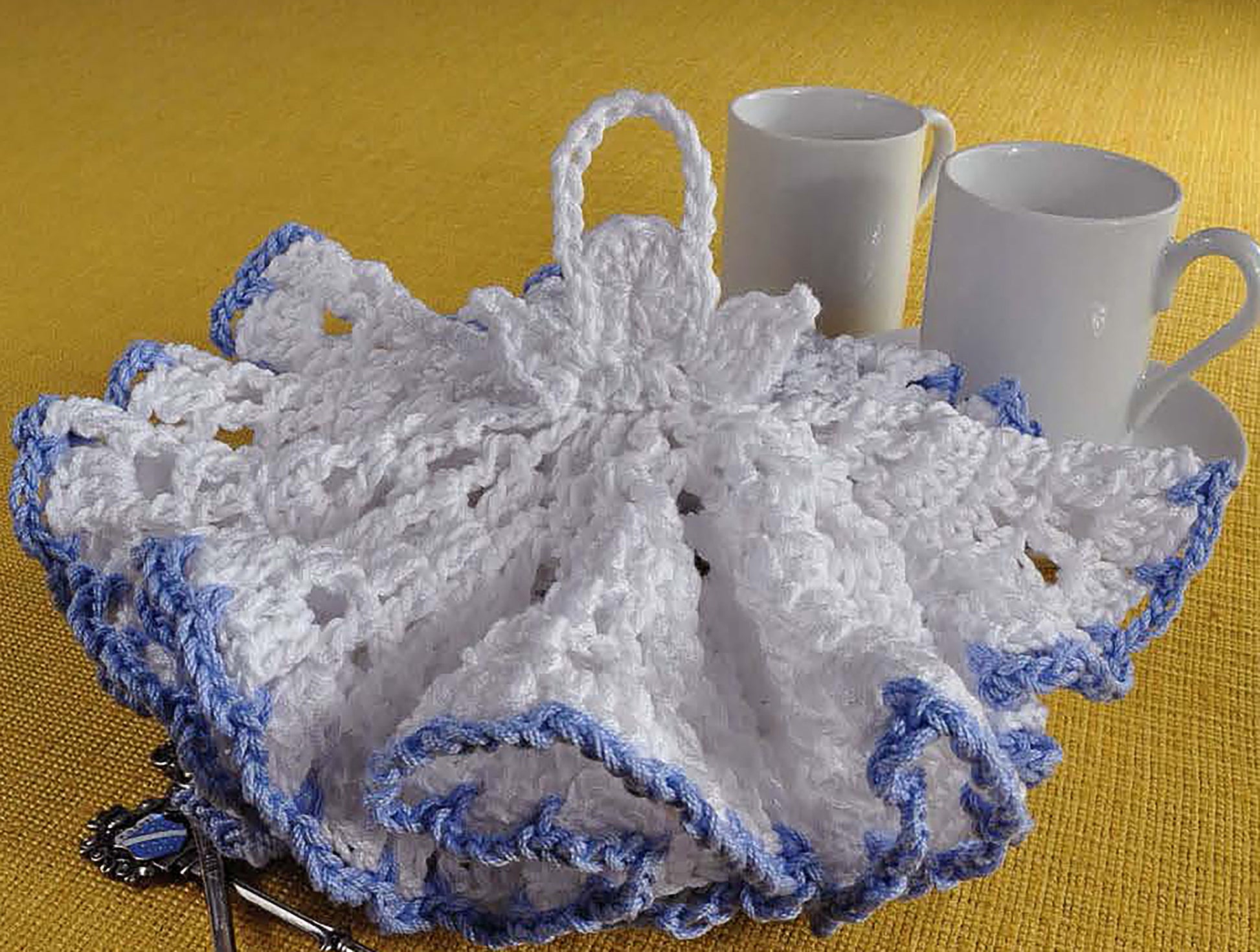 The "Complete Guide to Symbol Crochet" by Leisure Arts features a white, dress-shaped doily with blue edging—showing off intricate stitches, ideal for beginners and displayed beside two mugs and a spoon for project inspiration.