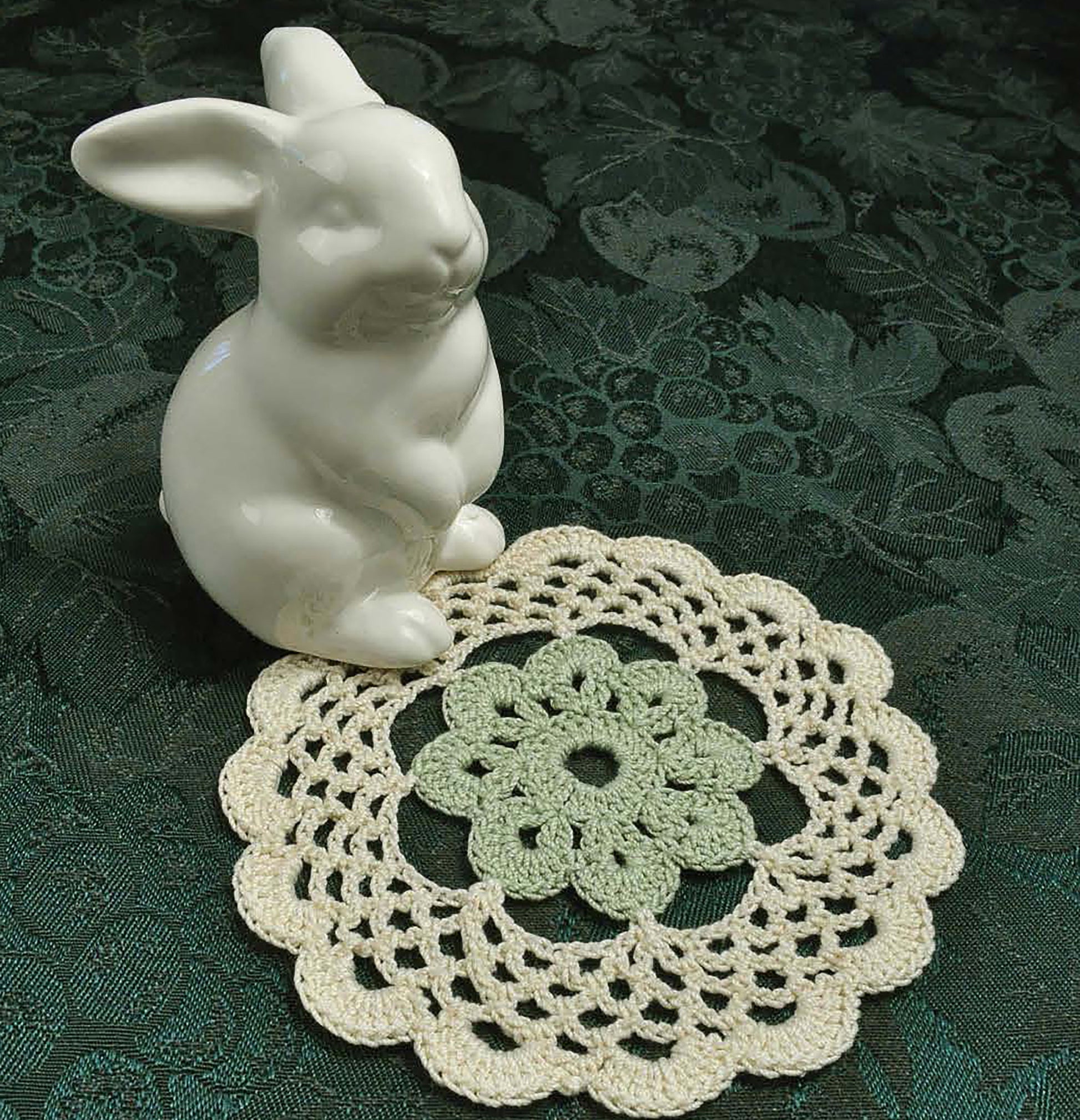 A white ceramic rabbit rests on dark green fabric beside a handmade doily with beautiful stitches—an inviting accent for crochet fans looking to start new projects using the Complete Guide to Symbol Crochet from Leisure Arts.