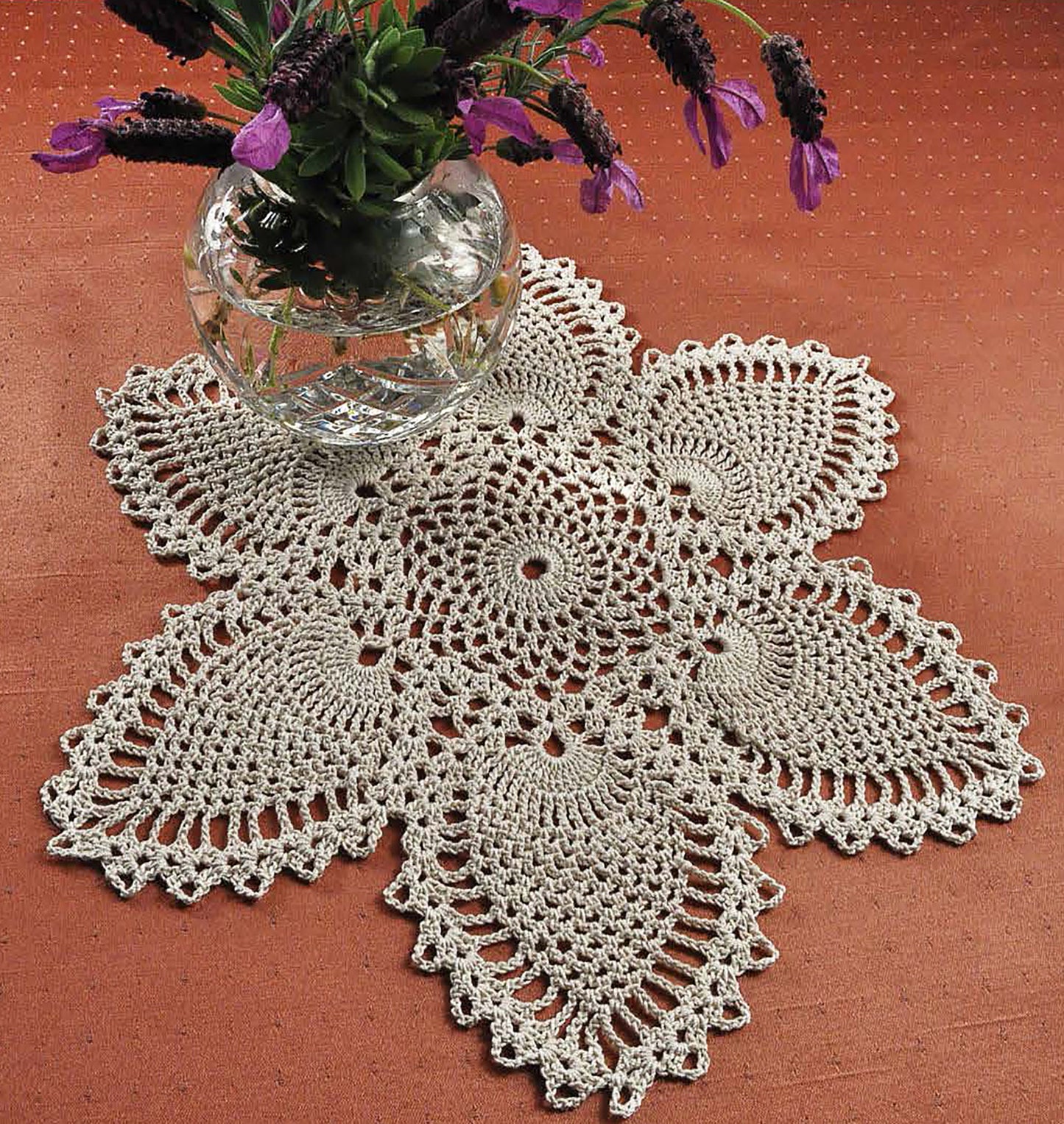 A round vase with purple flowers sits on a cream crocheted doily featuring six-petal stitches, inspired by patterns in Leisure Arts' Complete Guide to Symbol Crochet, displayed on a rust-colored surface.