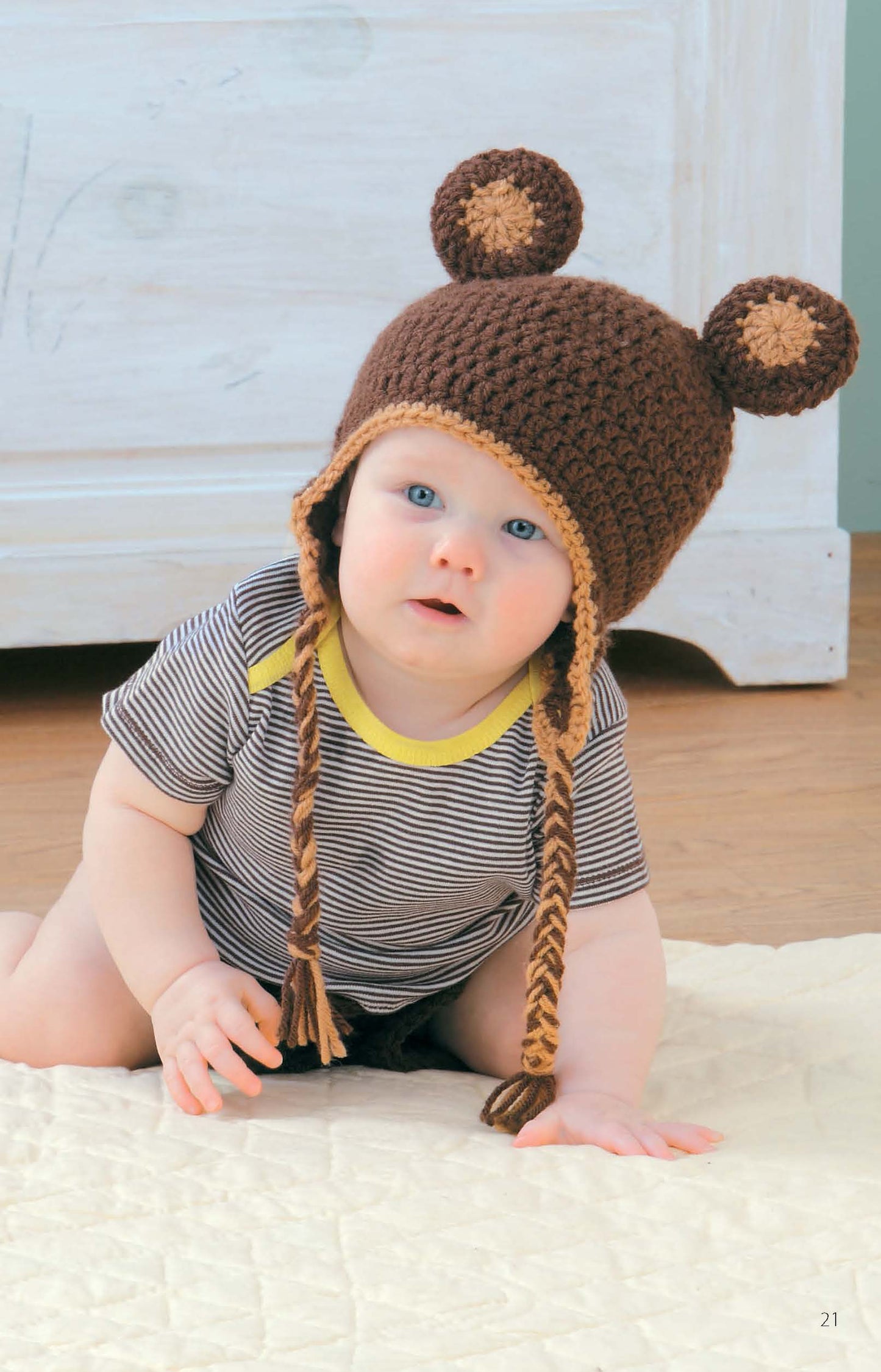 A baby wearing a brown crocheted bear hat with ear flaps from Leisure Arts' "Hats & Diaper Covers Book 2" digital download sits on a white blanket in a striped onesie.