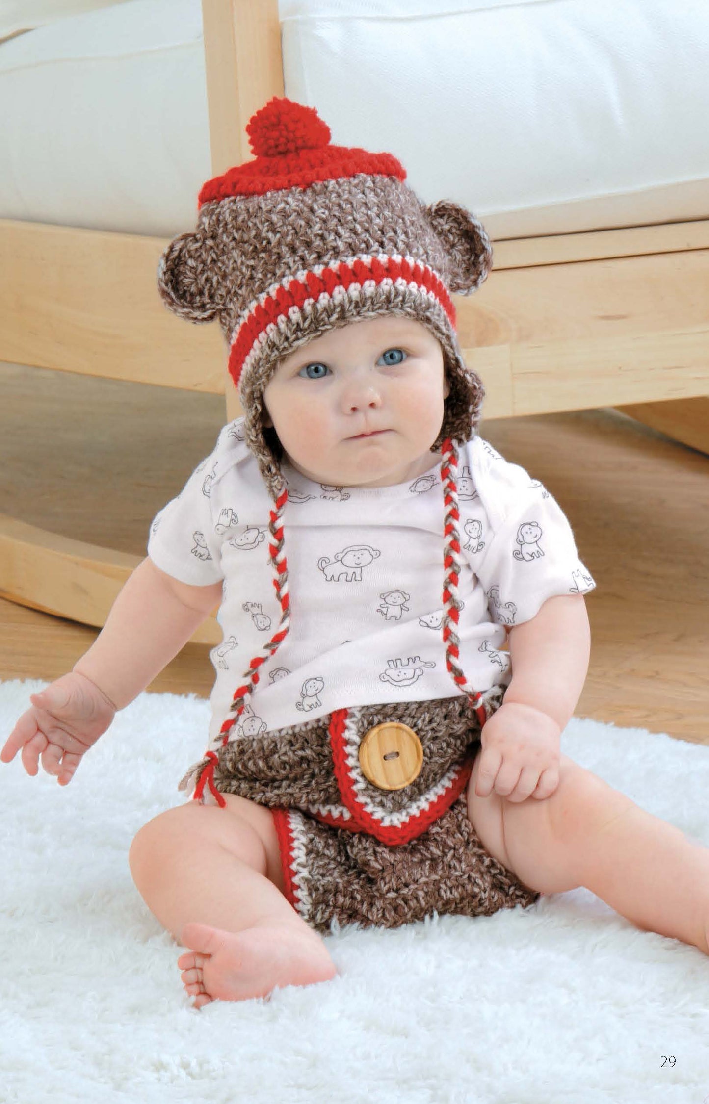 A baby wears a brown and red crochet set with bear ears and a pom-pom, made from the "Hats & Diaper Covers Book 2 Digital Download" by Leisure Arts, sitting on a white rug with a wooden chair in the background.