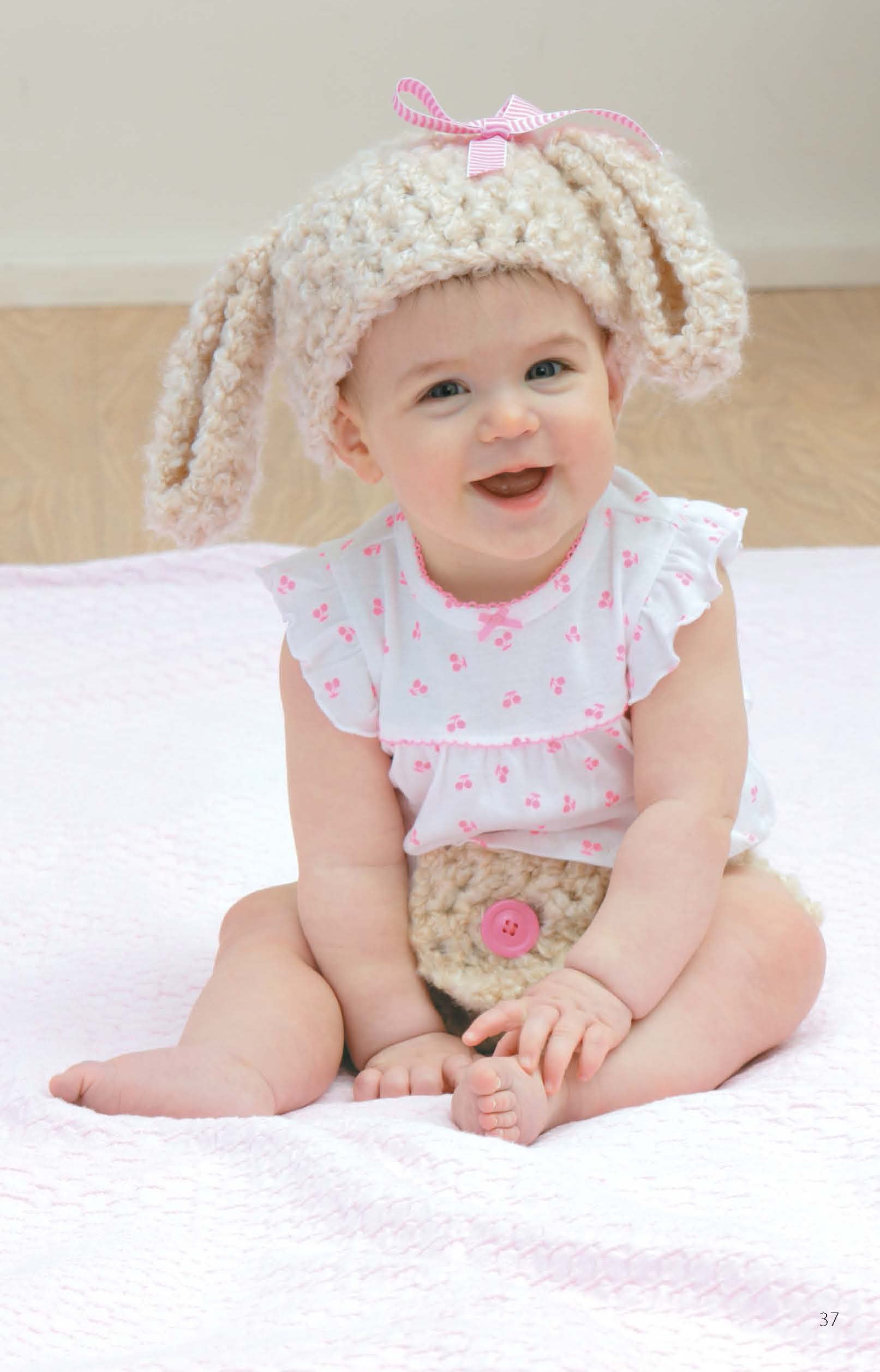 A smiling baby wears a cute bunny hat and diaper cover made from crochet patterns in the "Hats & Diaper Covers Book 2 Digital Download" by Leisure Arts, sitting on a pink blanket in a floral white outfit.