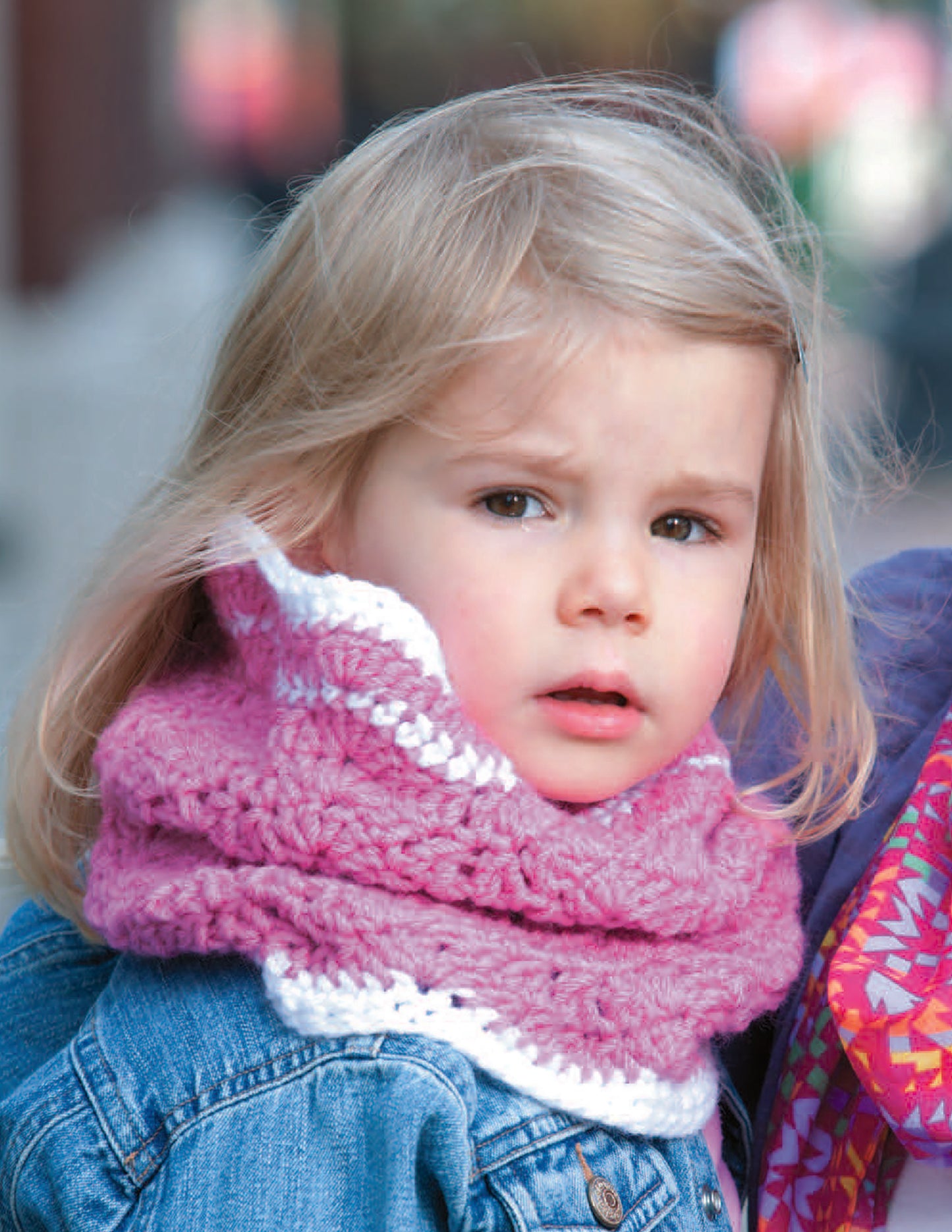 A young child with blonde hair wears a pink and white scarf made using patterns from Leisure Arts’ Crochet Celebrity Baby Fashion Digital Download, paired with a denim jacket and looking to the side with a neutral expression.