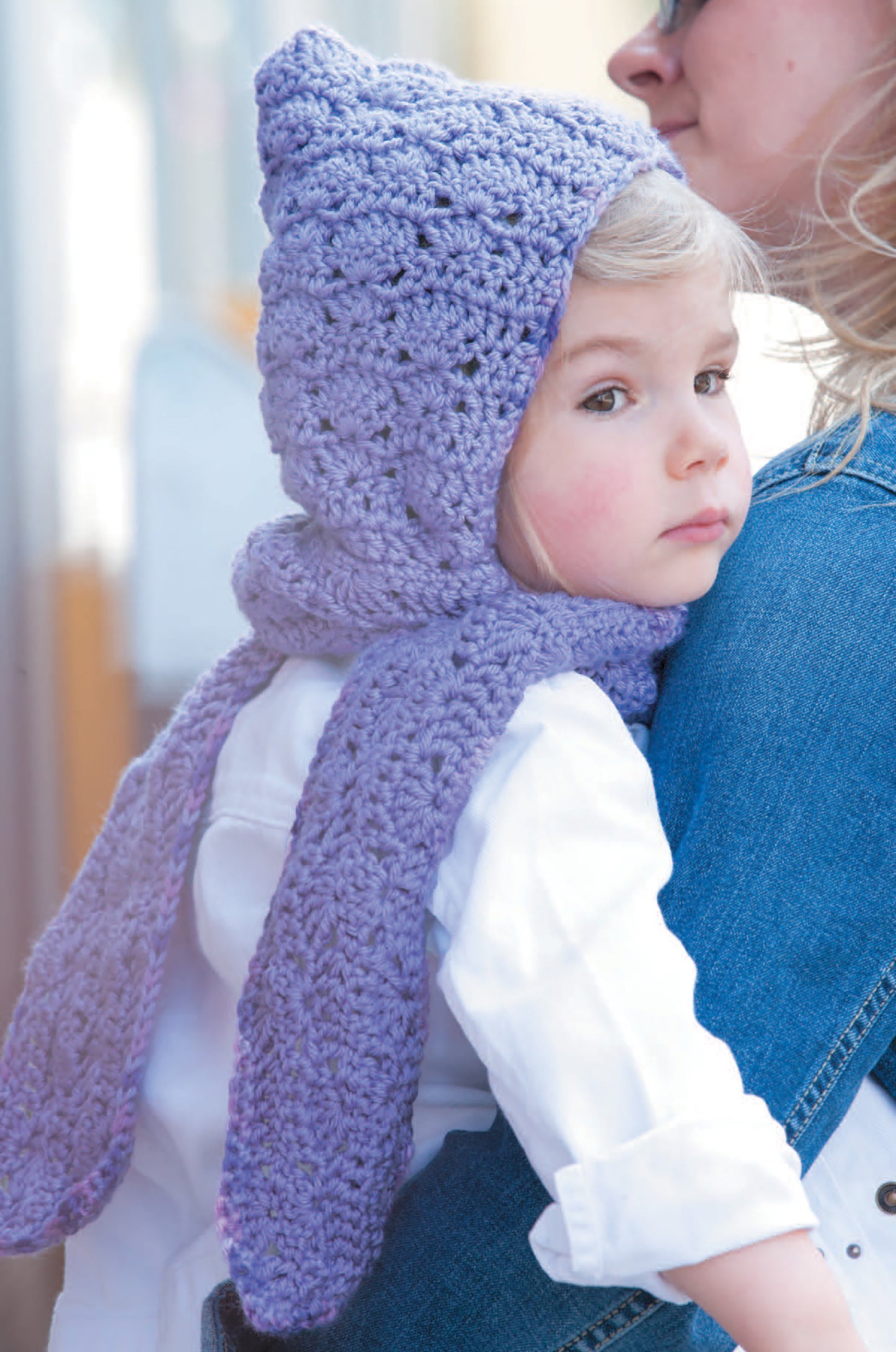 A young child in a purple hooded scarf—featured in the Crochet Celebrity Baby Fashion Digital Download by Leisure Arts—is held by an adult in a denim jacket, calmly looking over their shoulder.
