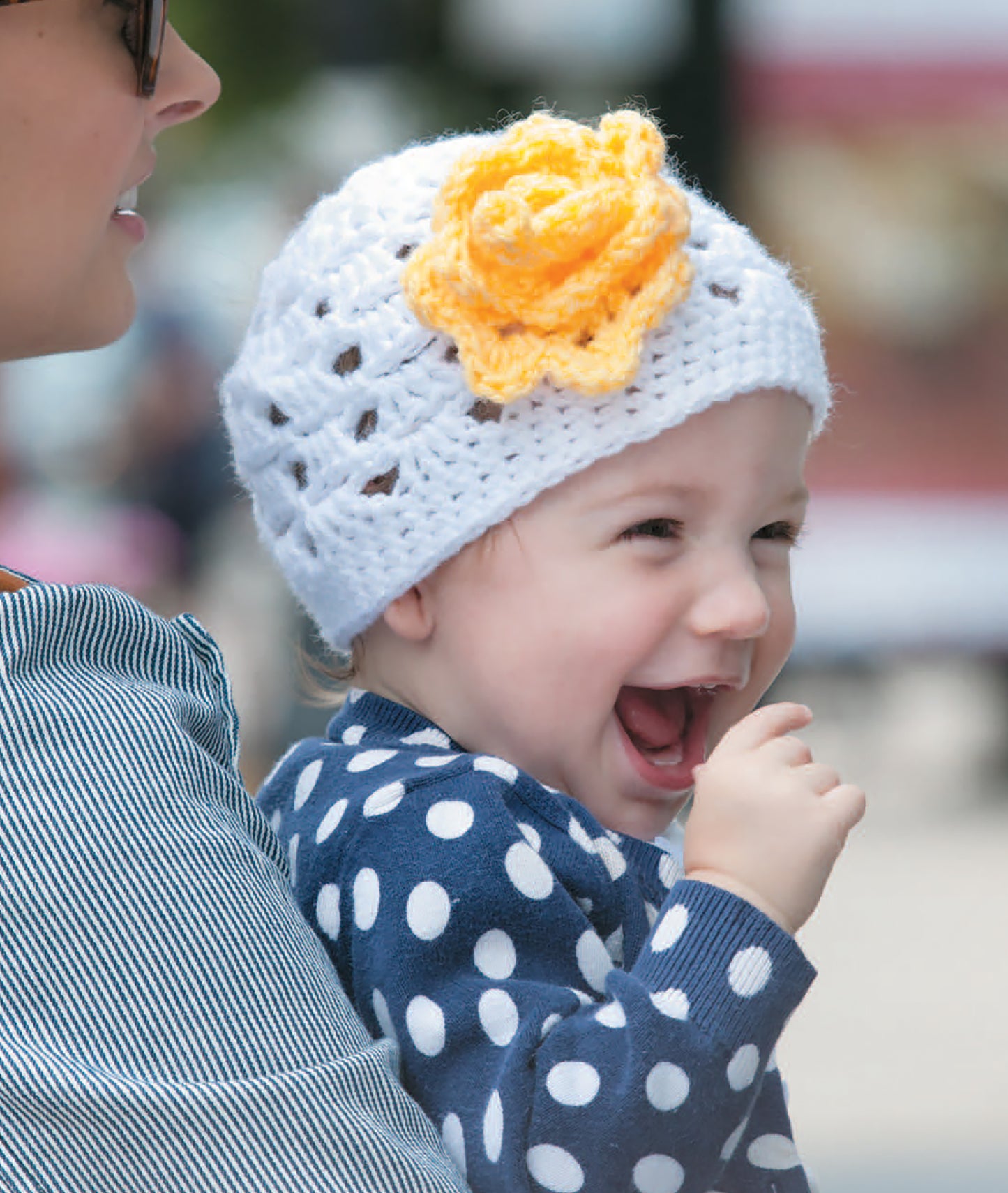 A smiling baby in a white crochet hat with a yellow flower and navy polka-dot sweater inspires creative projects—find more ideas in the Crochet Celebrity Baby Fashion Digital Download by Leisure Arts.