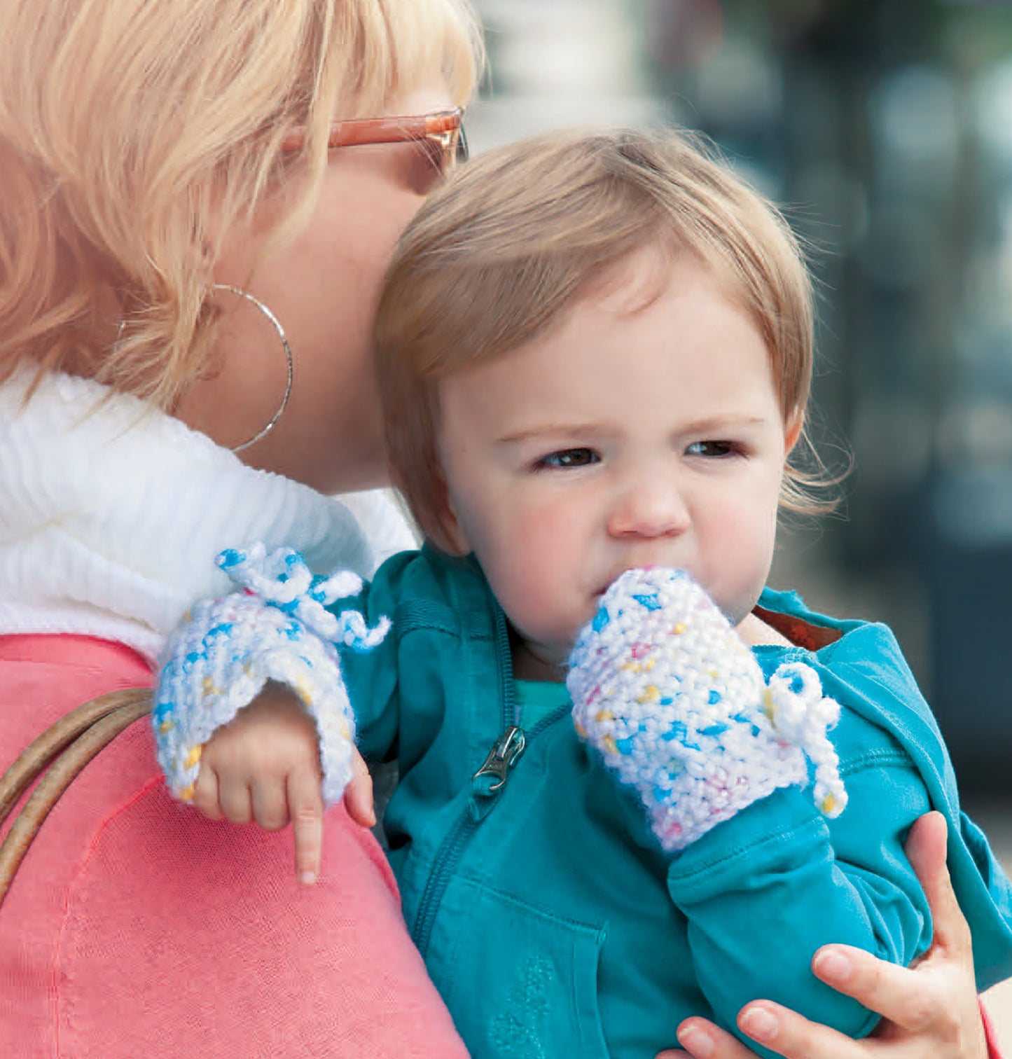 A woman holds a young child in a teal jacket and colorful knit mittens, inspiring baby crochet designs like those in Leisure Arts’ Crochet Celebrity Baby Fashion Digital Download. The smiling woman wears glasses, hoop earrings, and has short blonde hair.