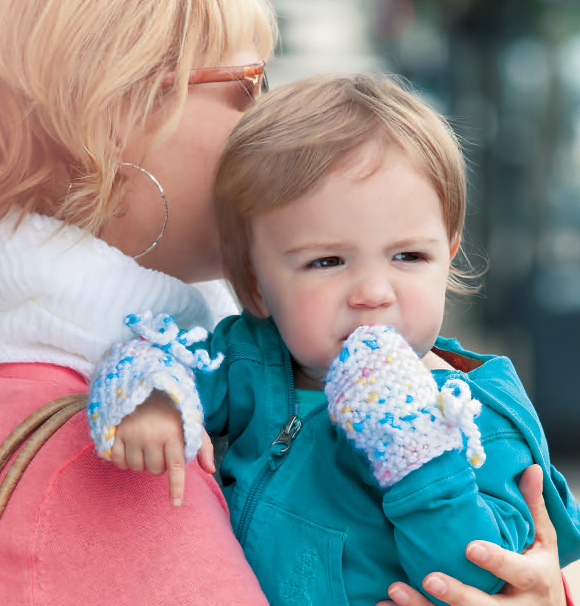 A woman holds a young child in a teal jacket and colorful knit mittens, inspiring baby crochet designs like those in Leisure Arts’ Crochet Celebrity Baby Fashion Digital Download. The smiling woman wears glasses, hoop earrings, and has short blonde hair.