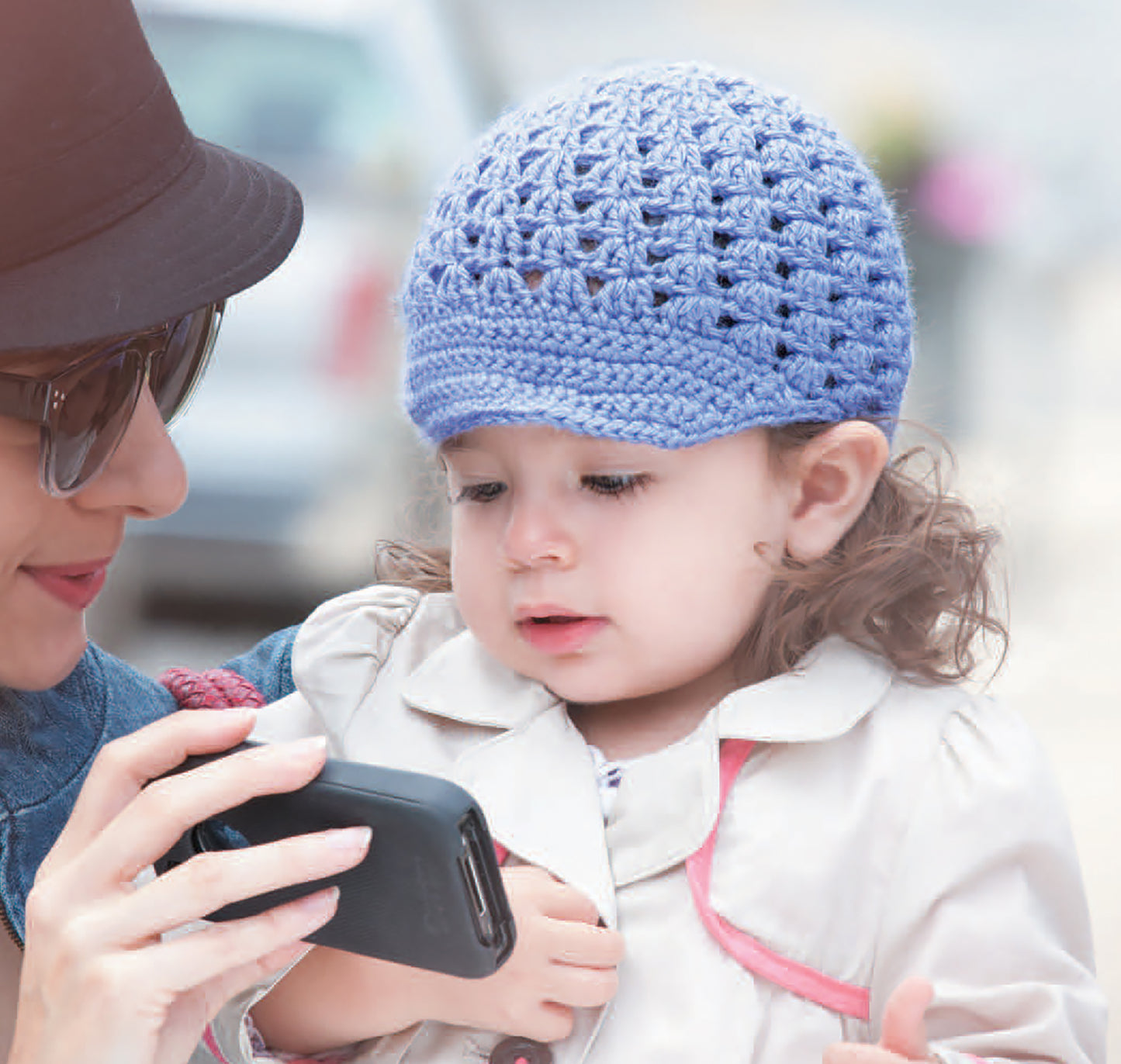 A woman in sunglasses shows a smartphone to a young child in a purple crocheted hat and light coat, featuring Crochet Celebrity Baby Fashion Digital Download by Leisure Arts, as the child sits on her lap outdoors.