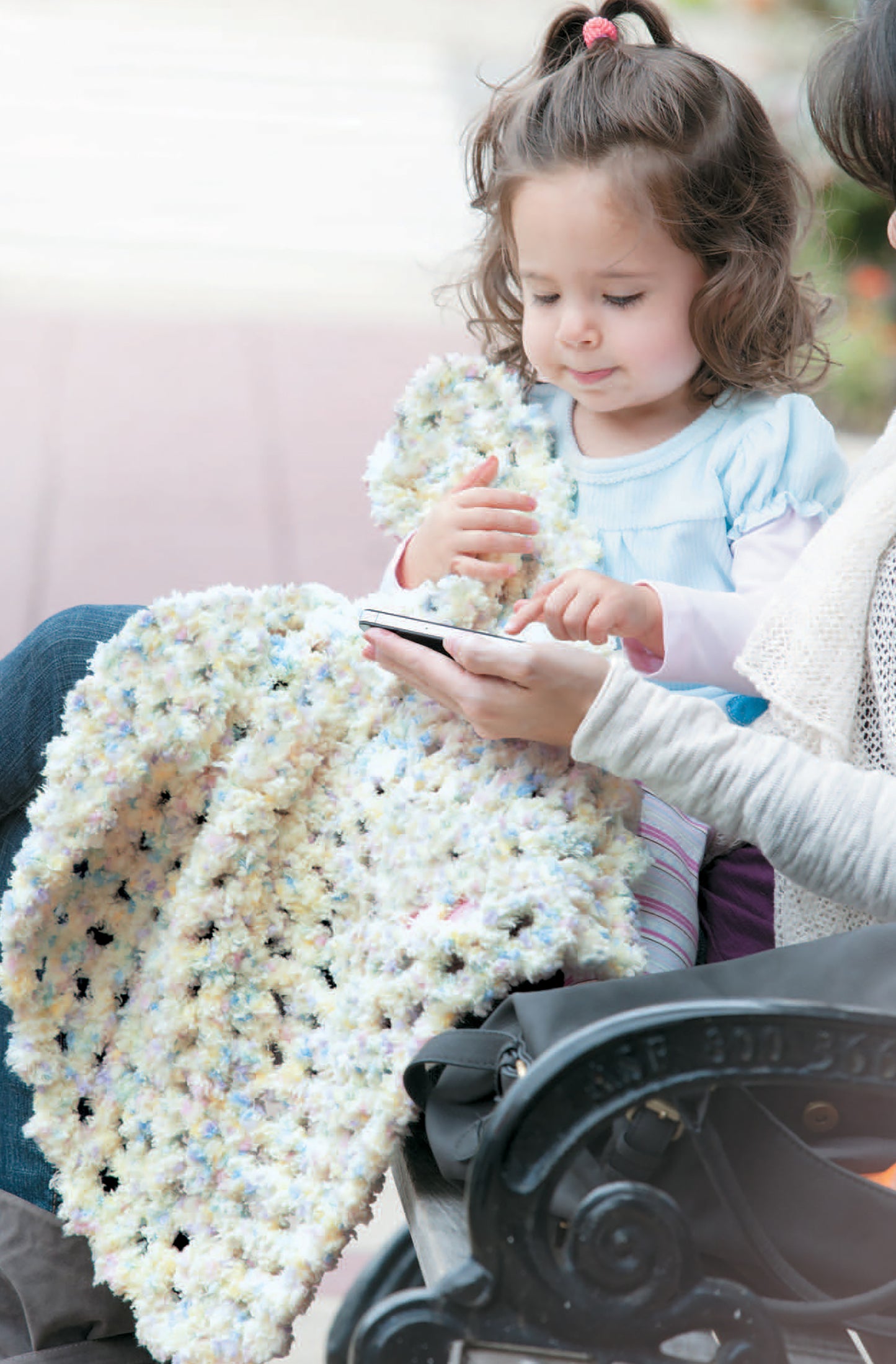 A young girl sits on a bench holding a pastel blanket made from the Crochet Celebrity Baby Fashion Digital Download by Leisure Arts, while an adult beside her uses a smartphone. The girl looks at the phone, appearing curious and engaged.