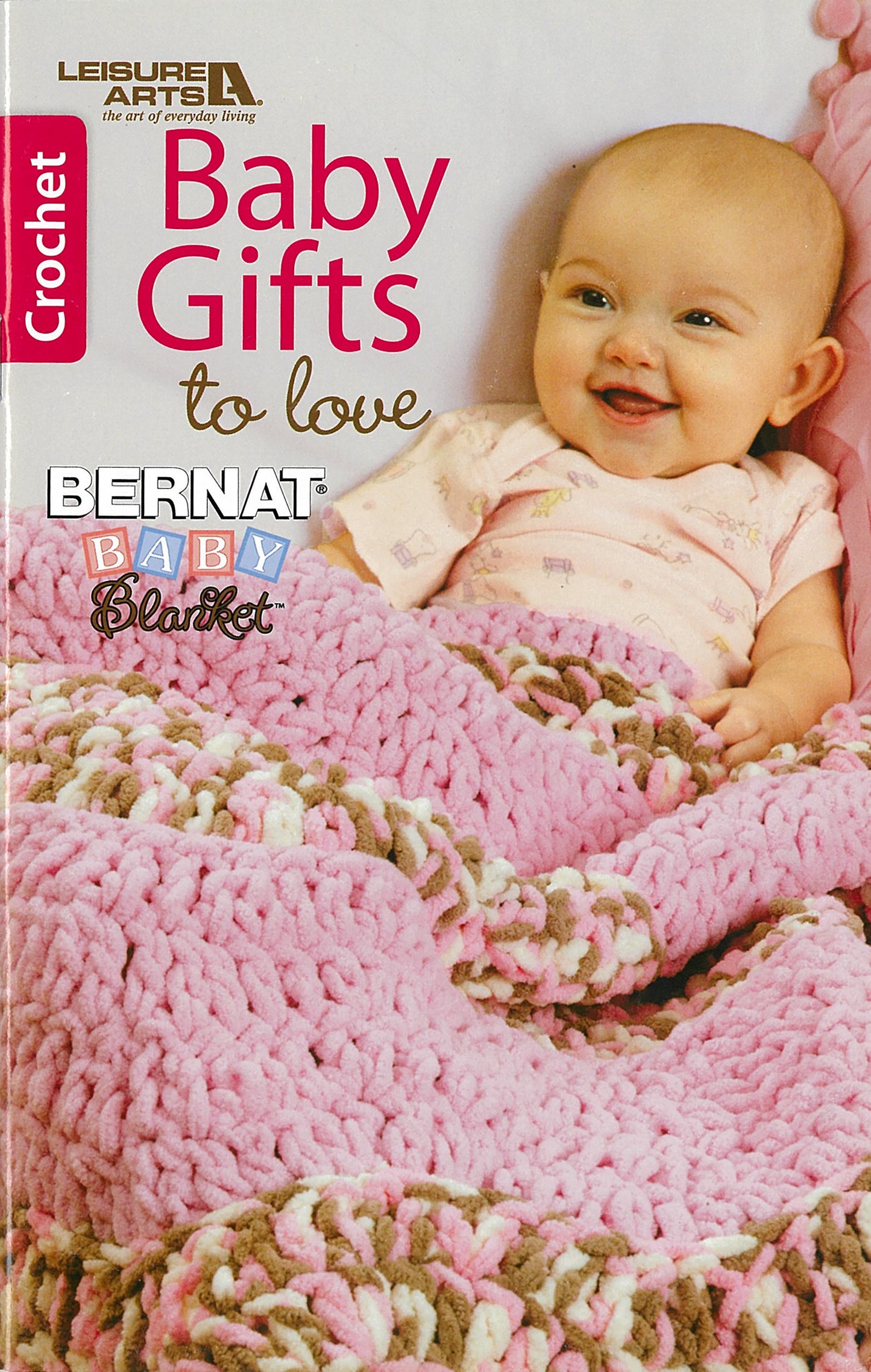 A smiling baby in pink lies under a chunky crocheted blanket. Beginner crafters will love "Baby Gifts to Love—Make Ultra-Soft Wraps, Hats & More!" by Leisure Arts. Print ISBN included.