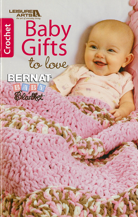 A smiling baby in pink lies under a chunky crocheted blanket. Beginner crafters will love "Baby Gifts to Love—Make Ultra-Soft Wraps, Hats & More!" by Leisure Arts. Print ISBN included.