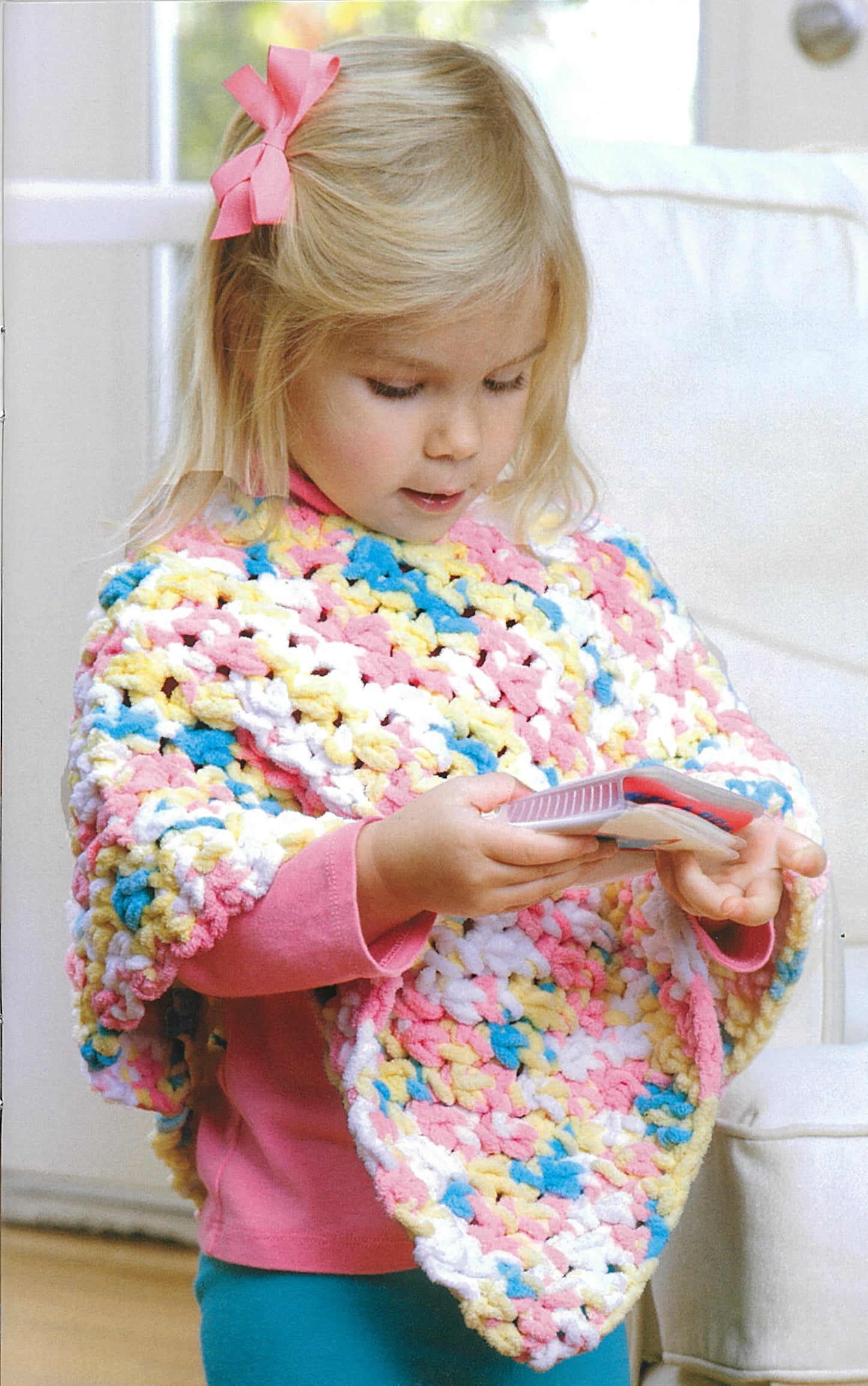 A young blonde girl with a pink bow wears a vibrant crocheted poncho from “Baby Gifts to Love—Make Ultra-Soft Wraps, Hats & More!” by Leisure Arts and holds a pink and white hairbrush while standing indoors near a white chair.