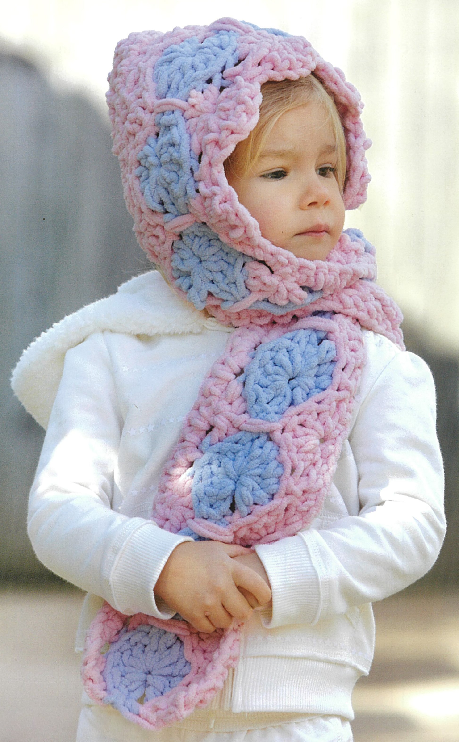 A young child wears a chunky crocheted hooded scarf with pink and blue hexagons over a white jacket—perfect inspiration for beginner projects in "Baby Gifts to Love—Make Ultra-Soft Wraps, Hats & More!" by Leisure Arts.