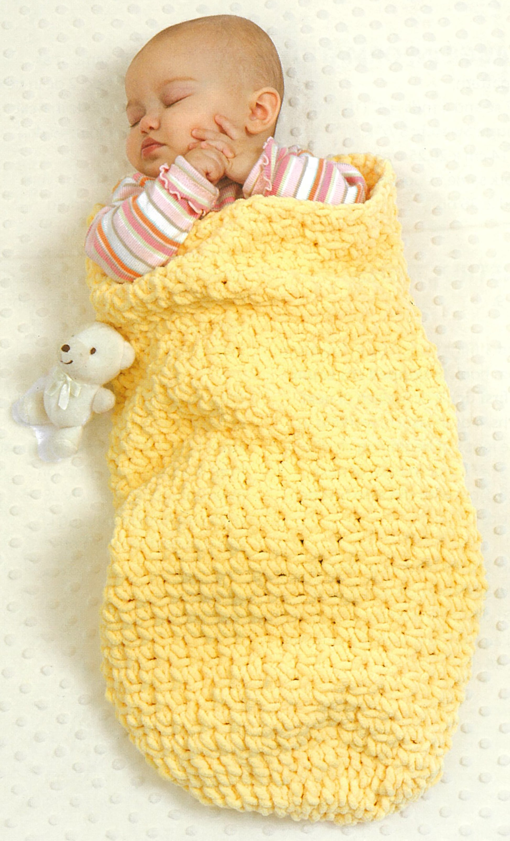 A sleeping baby wearing a pink striped outfit is wrapped in a yellow blanket made from "Baby Gifts to Love—Make Ultra-Soft Wraps, Hats & More!" by Leisure Arts, lying on a white surface with a small white teddy bear beside them.