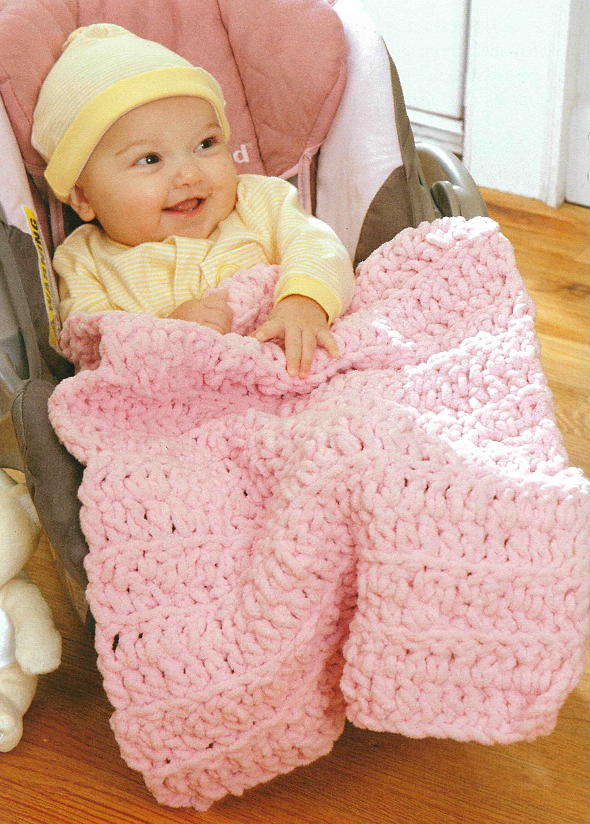 A smiling baby in a yellow hat and striped outfit sits in a car seat with a thick, soft pink crocheted blanket—an ideal beginner project from "Baby Gifts to Love—Make Ultra-Soft Wraps, Hats & More!" by Leisure Arts.