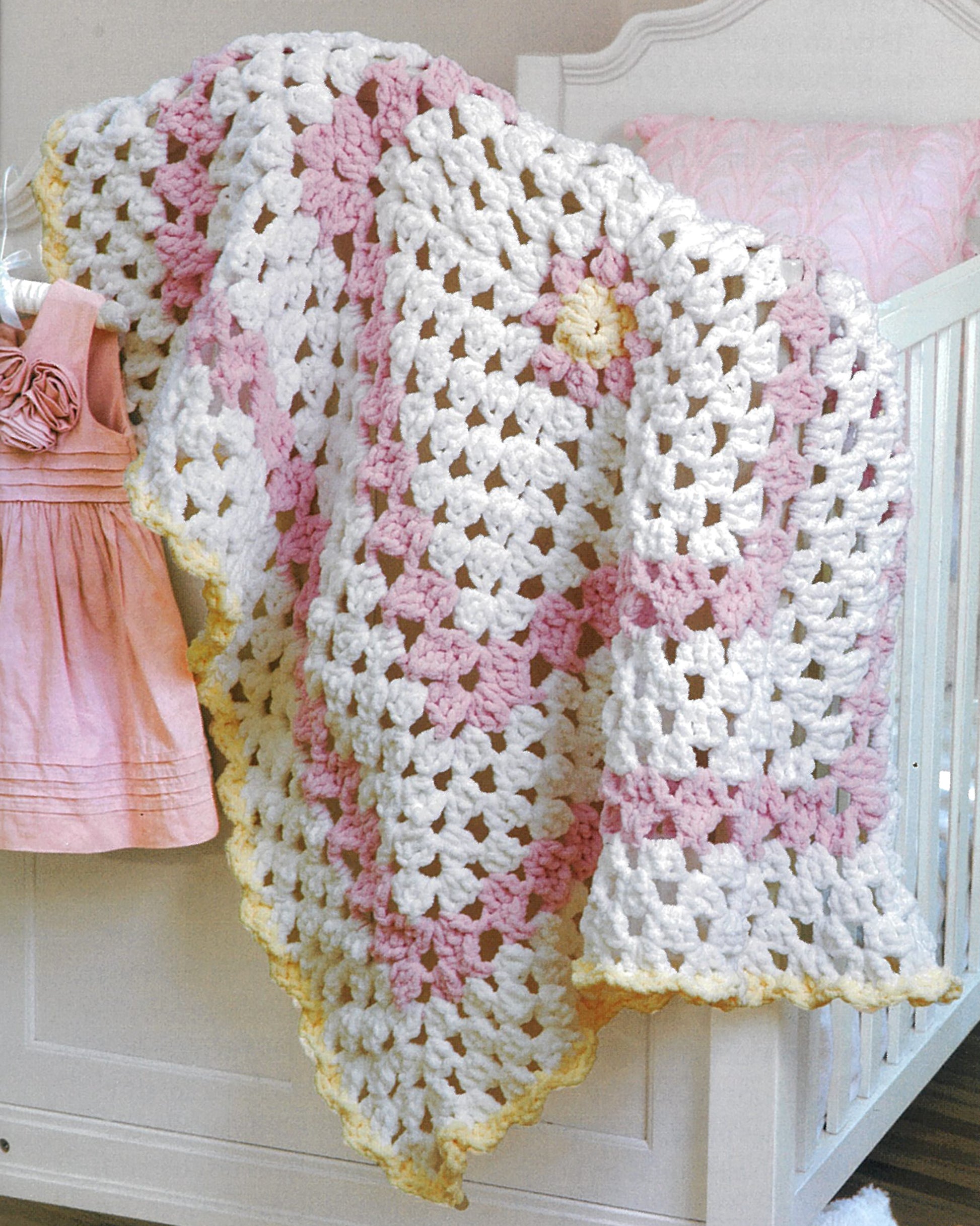A soft crocheted baby blanket with white, pink, and yellow granny squares—perfect for beginners—rests by a pink dress on a crib. Pattern from "Baby Gifts to Love—Make Ultra-Soft Wraps, Hats & More!" by Leisure Arts.