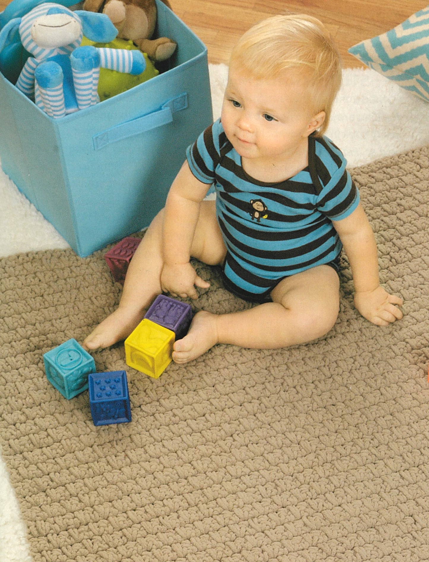 A blonde baby in a blue and black striped onesie sits on a beige rug playing with colorful blocks—the ideal setting to gift creations from "Baby Gifts to Love—Make Ultra-Soft Wraps, Hats & More!" by Leisure Arts. A blue box of stuffed animals adds charm.