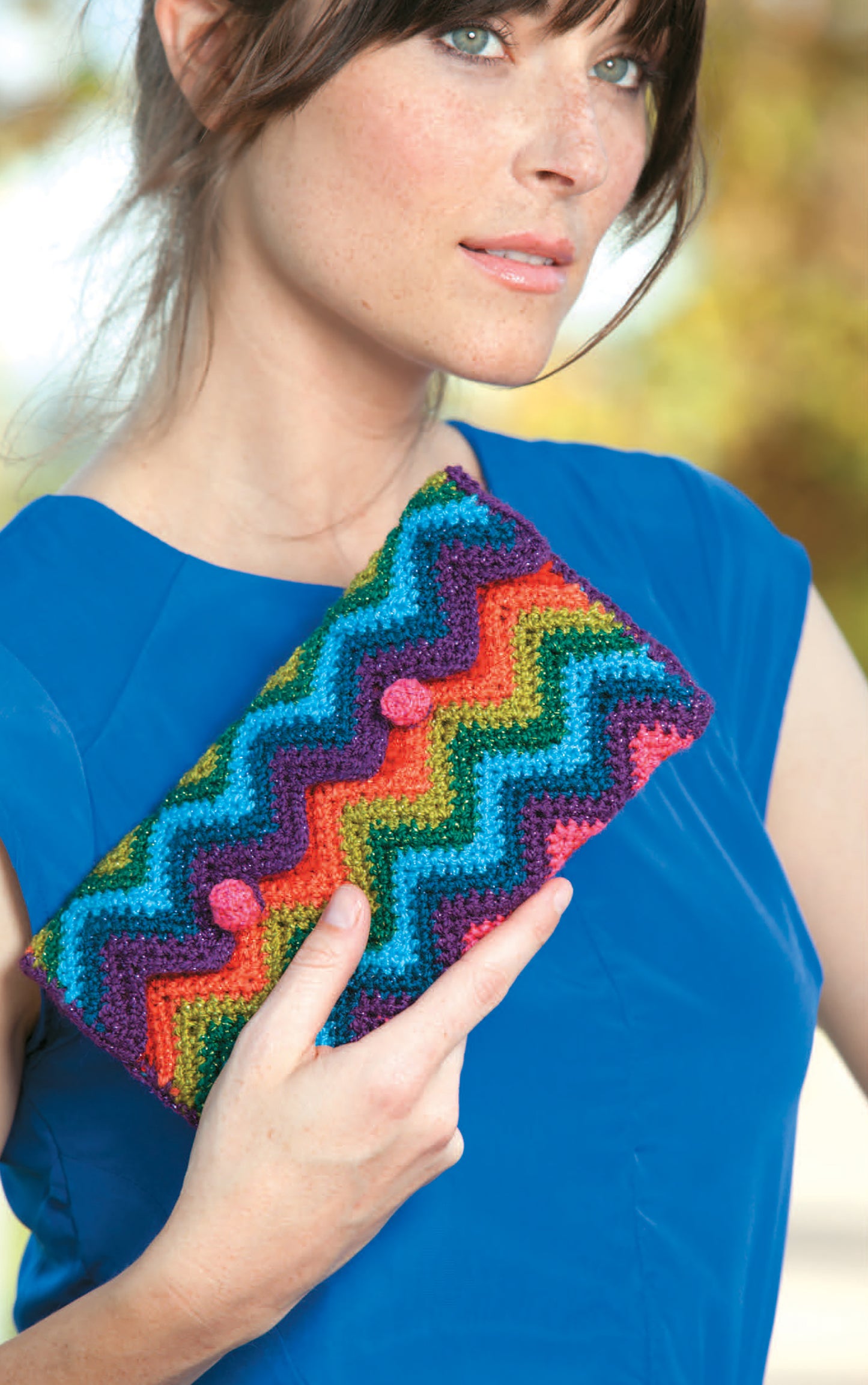 A woman in a blue dress holds a colorful, zigzag-patterned clutch made with Quick Itty Bitties Crochet by Leisure Arts, perfect for beginners, against a blurred outdoor background.