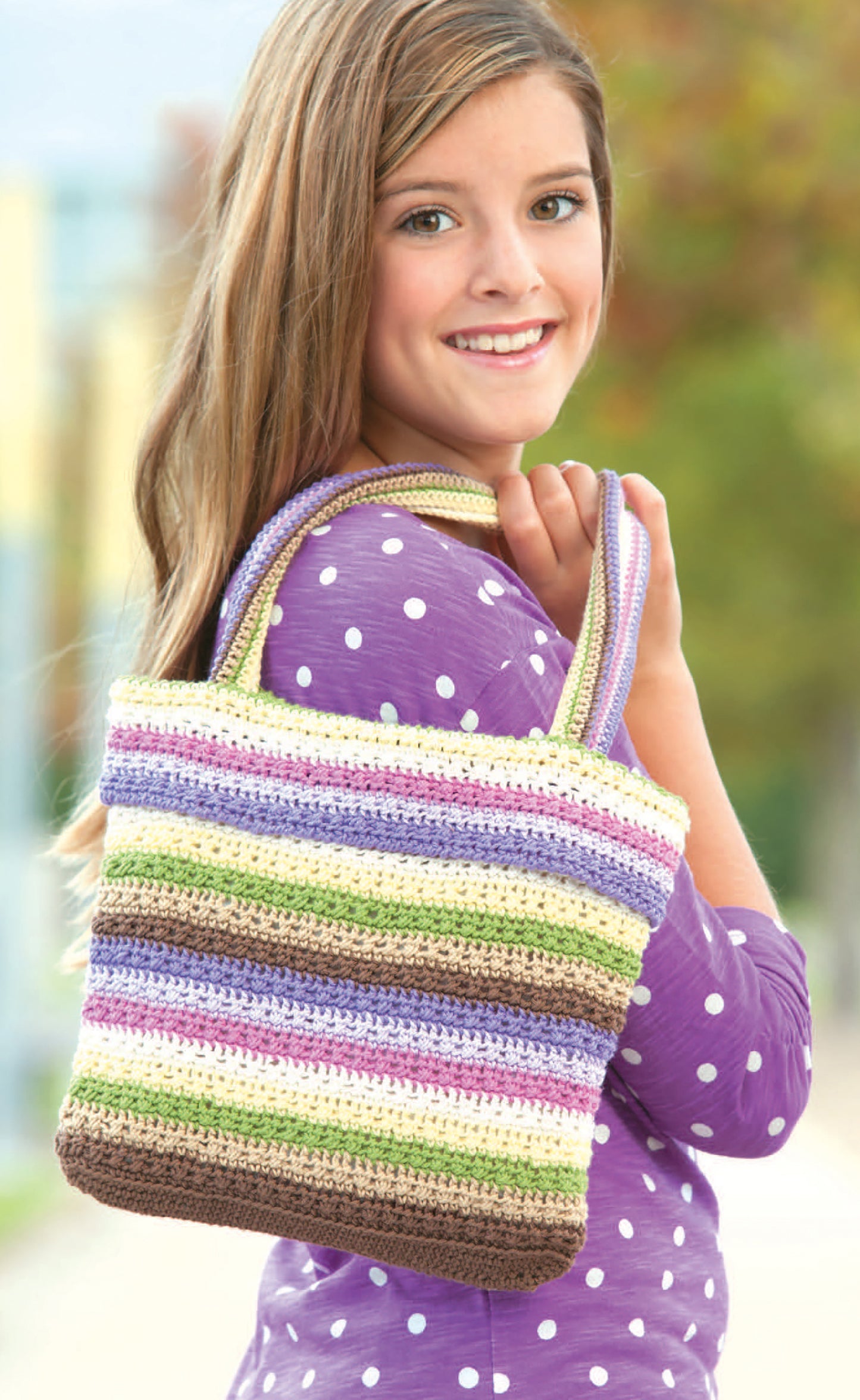 A smiling girl in a polka dot top holds a large striped crochet tote bag made with Quick Itty Bitties Crochet by Leisure Arts. The softly blurred outdoor background highlights this charming beginner project.
