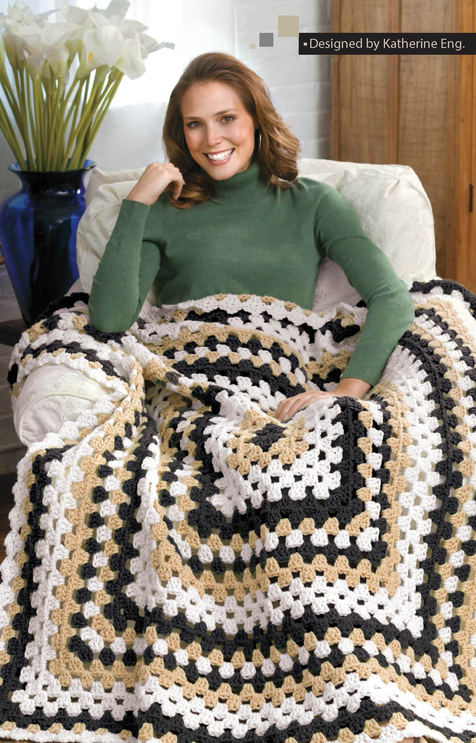A woman sits comfortably, smiling and wrapped in a large granny square blanket made from the “Crochet Throws Worked in the Round Digital Download” by Leisure Arts. A vase with white flowers sits in the background.