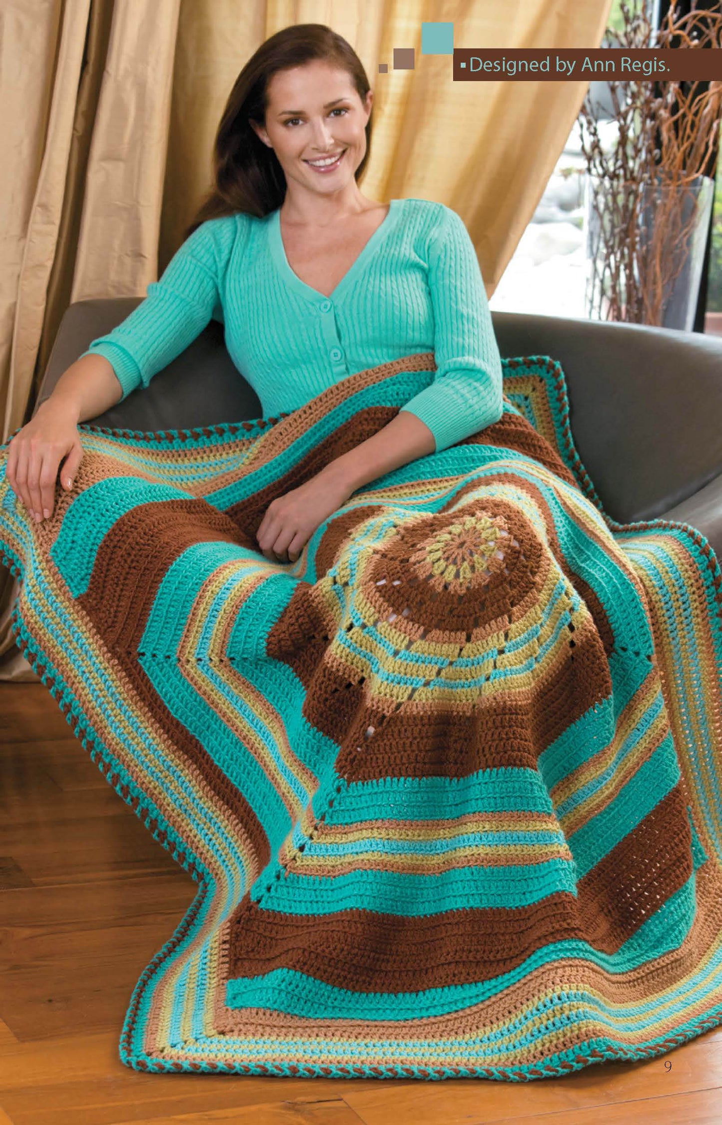 A woman smiles on a gray couch, wrapped in a colorful afghan made with Leisure Arts' "Crochet Throws Worked in the Round" digital pattern—ideal for easy to intermediate crochet enthusiasts.