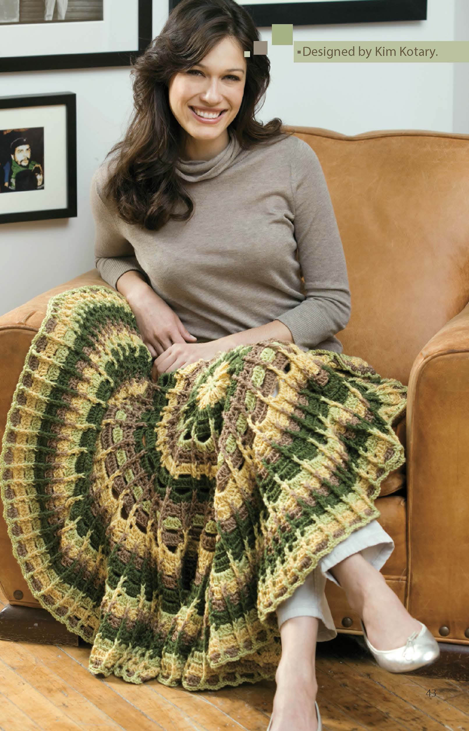 A woman sits on a tan couch with the Leisure Arts "Crochet Throws Worked in the Round Digital Download" afghan in tan, green, and brown draped over her lap. She's wearing a taupe turtleneck and light pants, with framed art hanging behind her.