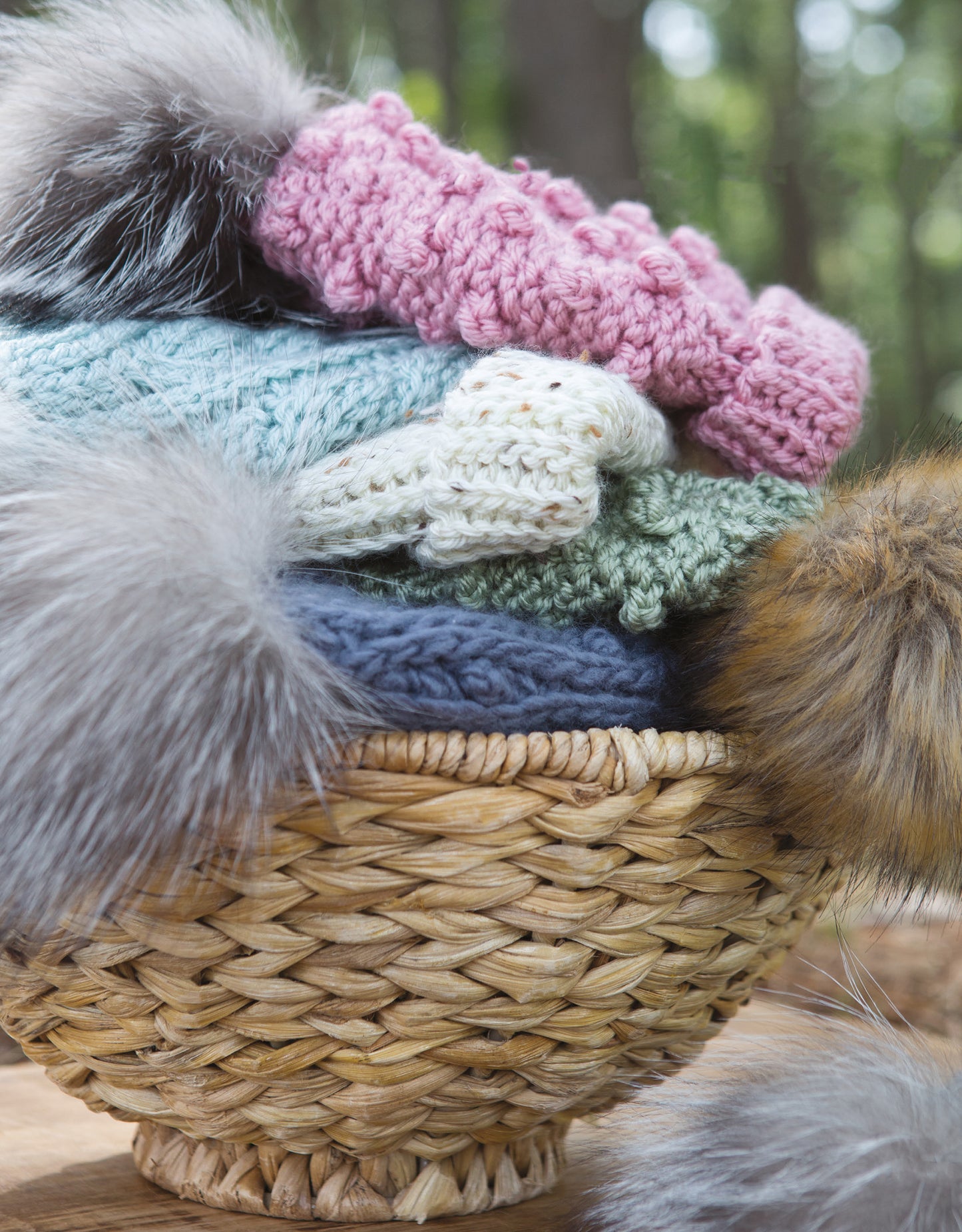 A woven basket displays a stack of colorful Alaskan Hats Using Slip Stitch from Leisure Arts, each with a fluffy faux fur pom-pom, set before a blurred green outdoor backdrop.