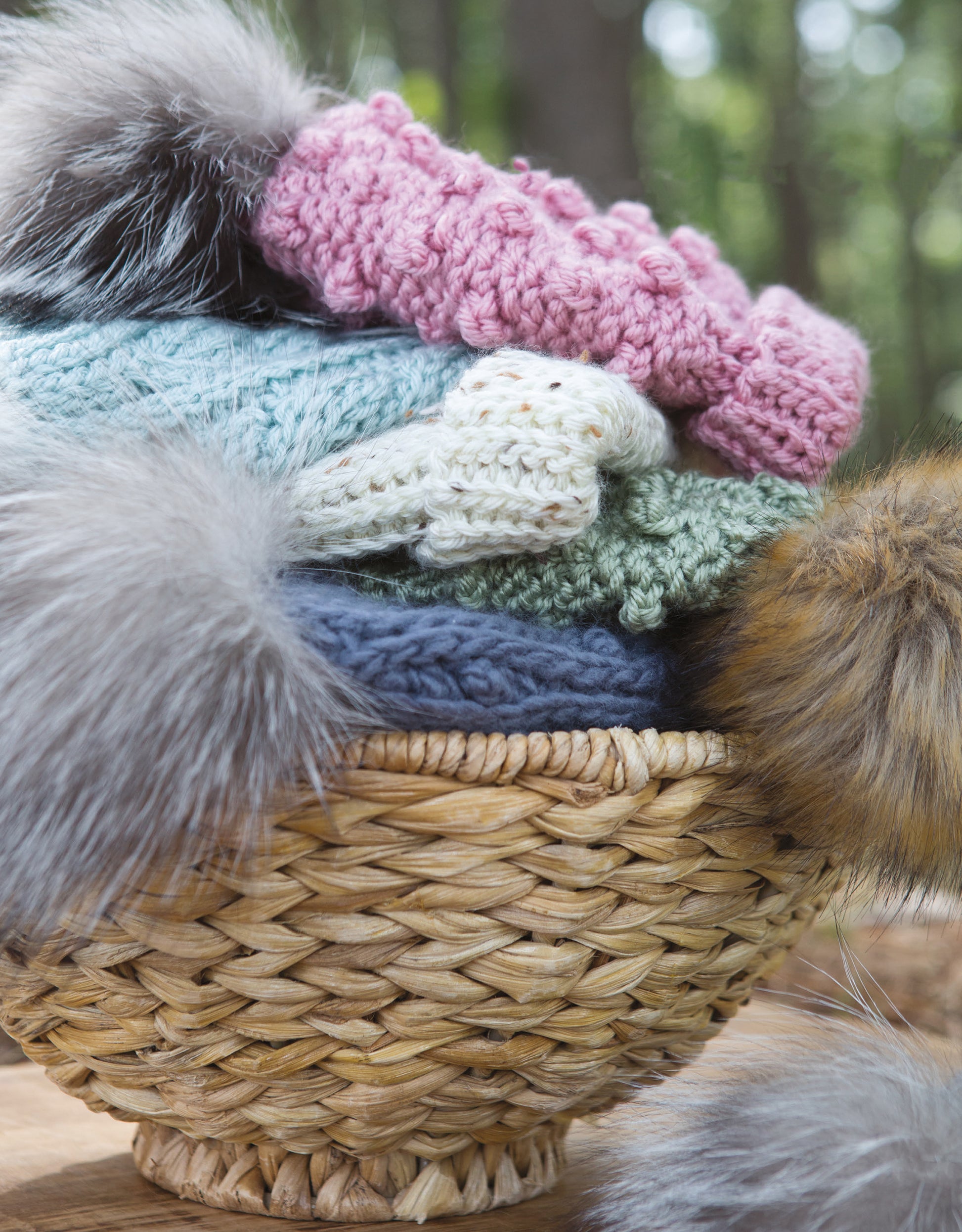 A woven basket displays a stack of colorful Alaskan Hats Using Slip Stitch from Leisure Arts, each with a fluffy faux fur pom-pom, set before a blurred green outdoor backdrop.