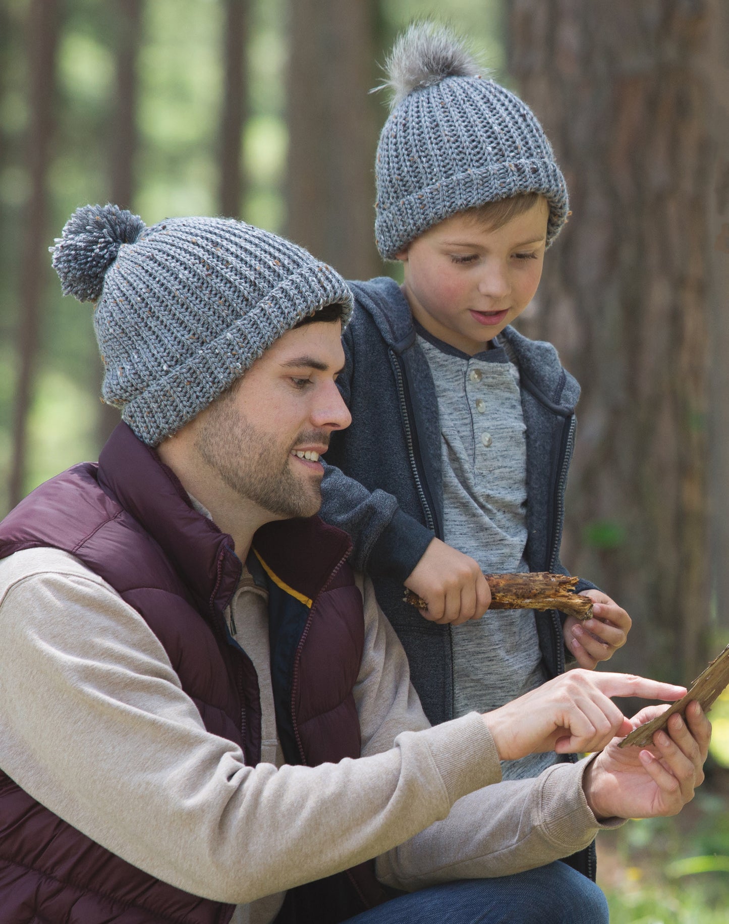 A man and a boy wear matching gray Alaskan Hats, made with the Leisure Arts Slip Stitch digital pattern, while exploring together in a forest. The man points at wood as the boy watches, both looking interested.