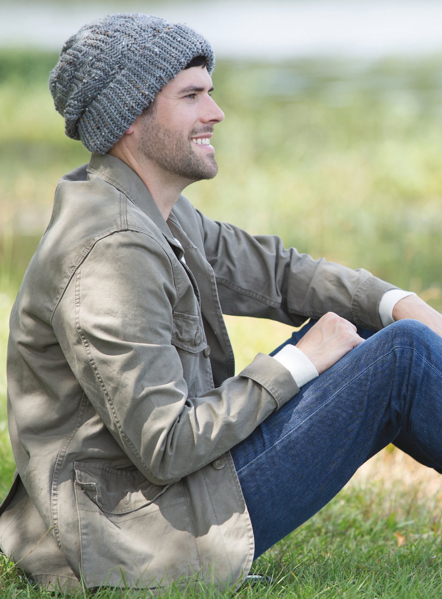 A man in a gray knit beanie, khaki jacket, and blue jeans smiles outdoors on grass. His beanie resembles the “Alaskan Hats Using Slip Stitch” digital pattern by Leisure Arts. Blurred greenery and water form the background.