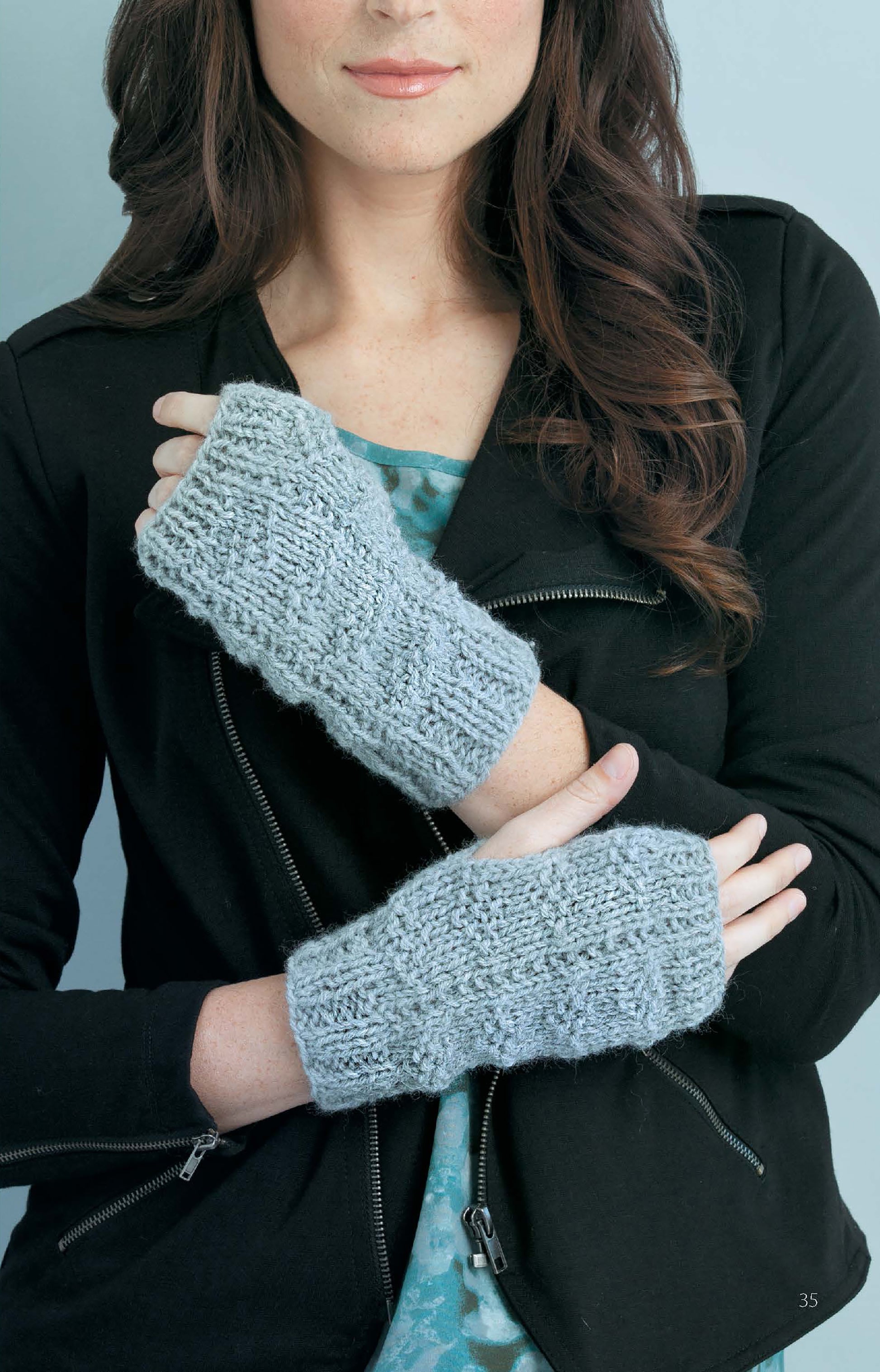 A woman with wavy brown hair models chunky light gray knit fingerless gloves, made using Leisure Arts’ “Learn to Knit—Easy Step-by-Step How-Tos, Eye-Catching Beginner Projects” digital download, against a pale blue background.