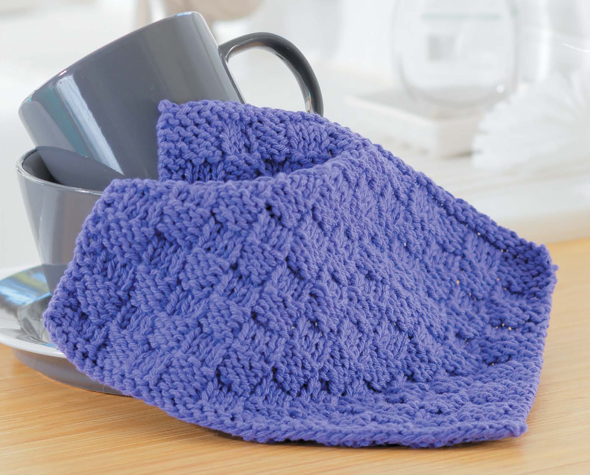 A purple, textured super bulky dishcloth from the Leisure Arts "Learn to Knit—Easy Step-by-Step How-Tos, Eye-Catching Beginner Projects" digital download is draped over gray mugs on wood, with a softly blurred white decor background.