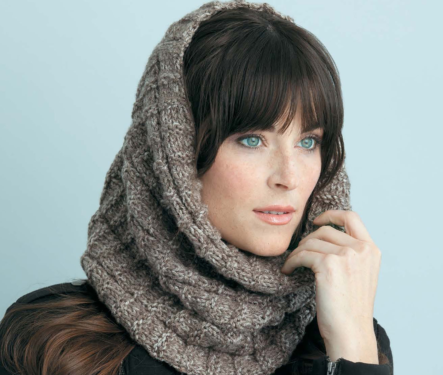 A woman with dark hair and blue eyes wears a chunky knit cowl from Learn to Knit—Easy Step-by-Step How-Tos, Eye-Catching Beginner Projects by Leisure Arts, paired with a black jacket against a light blue background.
