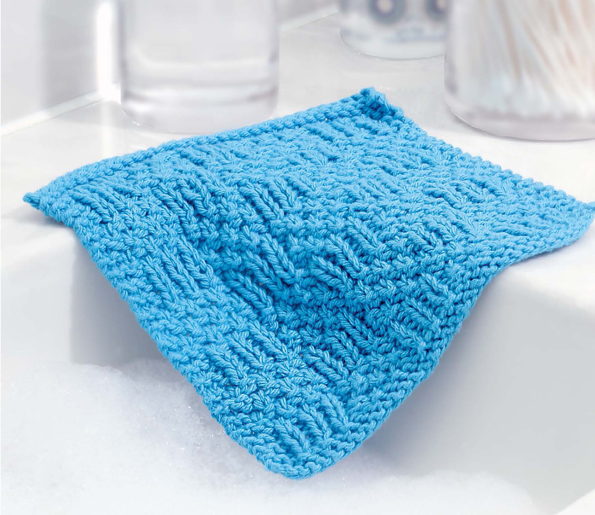 A digital download from Leisure Arts, "Learn to Knit—Easy Step-by-Step How-Tos, Eye-Catching Beginner Projects," rests by a white sink with soapy water, surrounded by blurred bathroom items in the background.