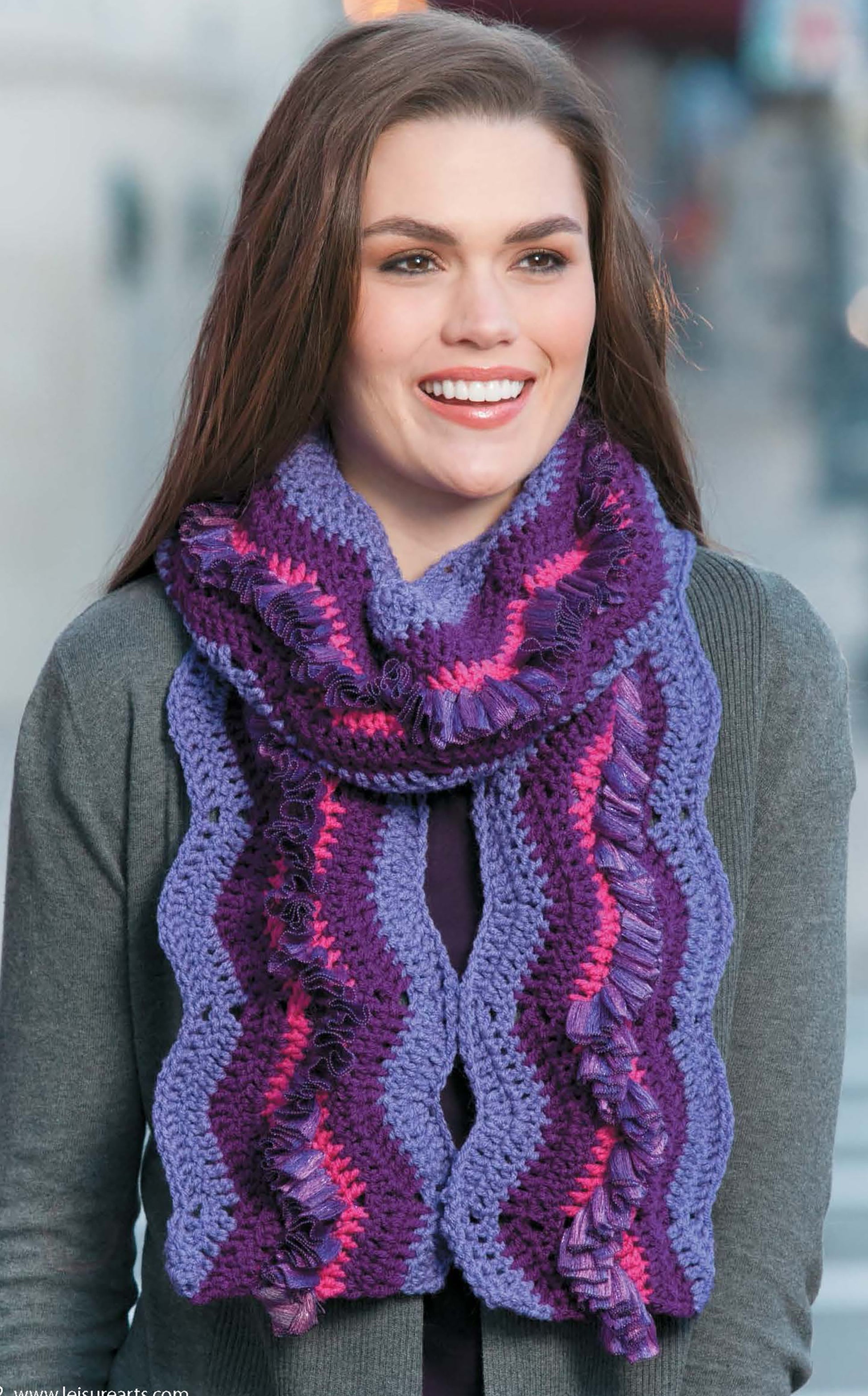 A smiling woman with long brown hair wears the Crochet Fashion Neckwear by Leisure Arts—a purple, pink, and lavender wavy-striped scarf—paired with a gray cardigan, standing on a city street.
