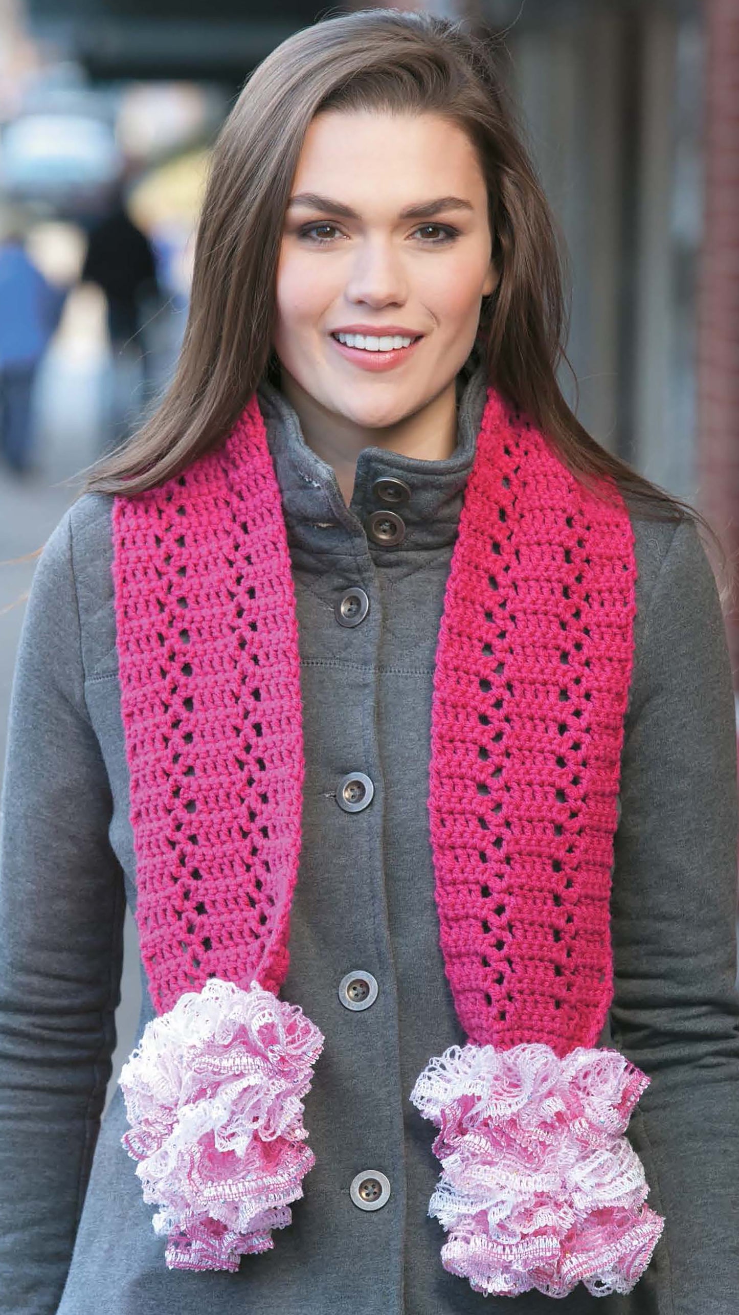 A woman stands outside on a sidewalk wearing the Crochet Fashion Neckwear by Leisure Arts—a gray buttoned coat paired with a bright pink, ruffled-edge crocheted scarf, perfect for scarf and neckwarmer enthusiasts.