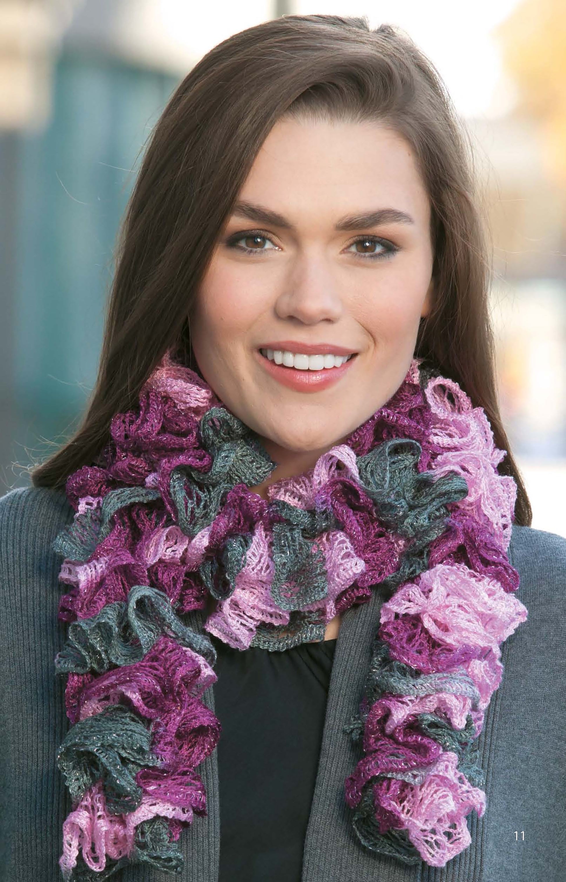A woman with long brown hair smiles at the camera, wearing a gray jacket and a ruffled, multicolored Crochet Fashion Neckwear scarf by Leisure Arts, standing outdoors in soft natural light.