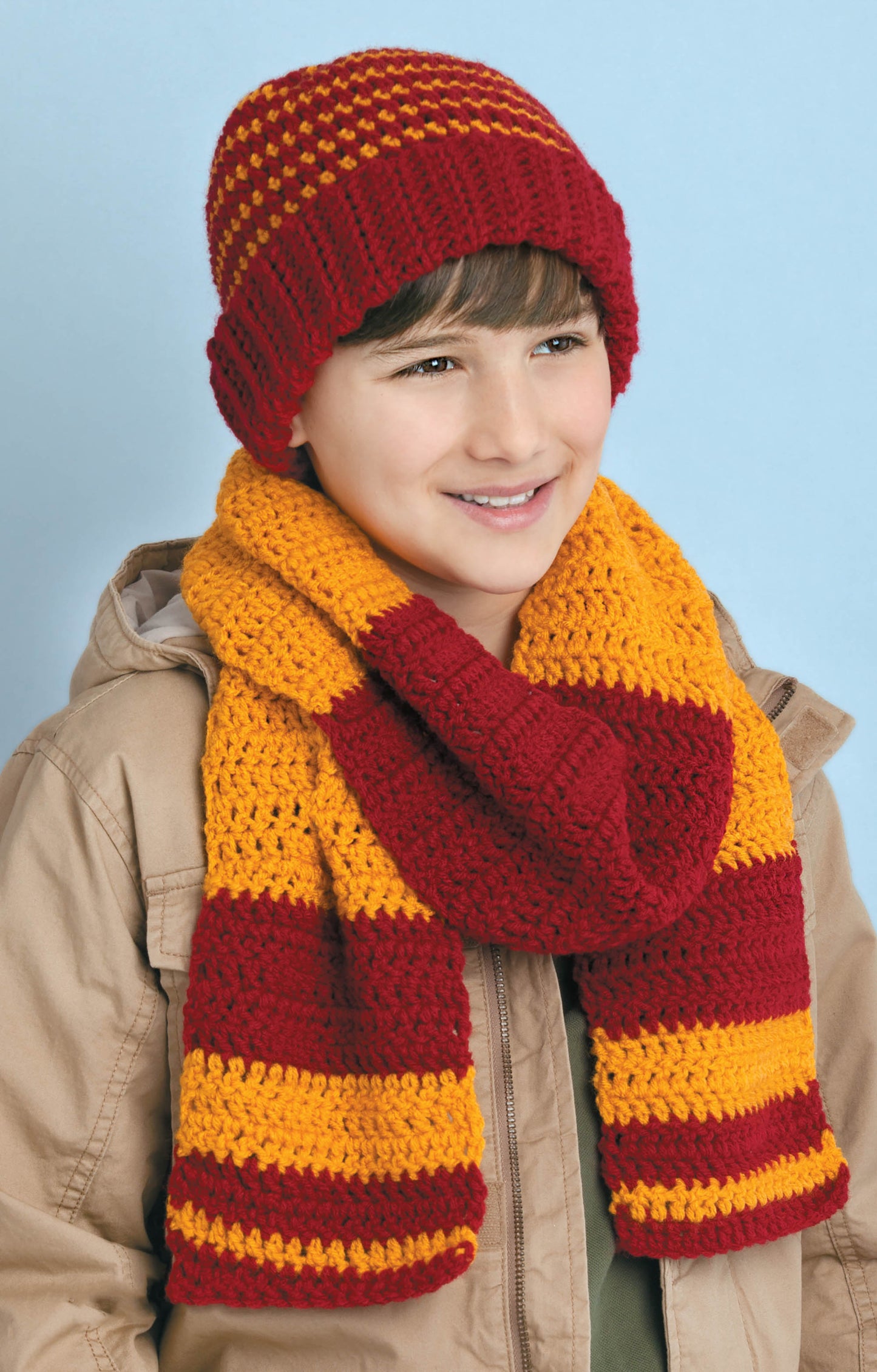 A smiling child in a red and gold striped knit hat and scarf set, shown against a light blue background. Find inspiration for cozy gifts from "Hats & Scarves for the Family" by Leisure Arts—perfect for winter crochet projects.
