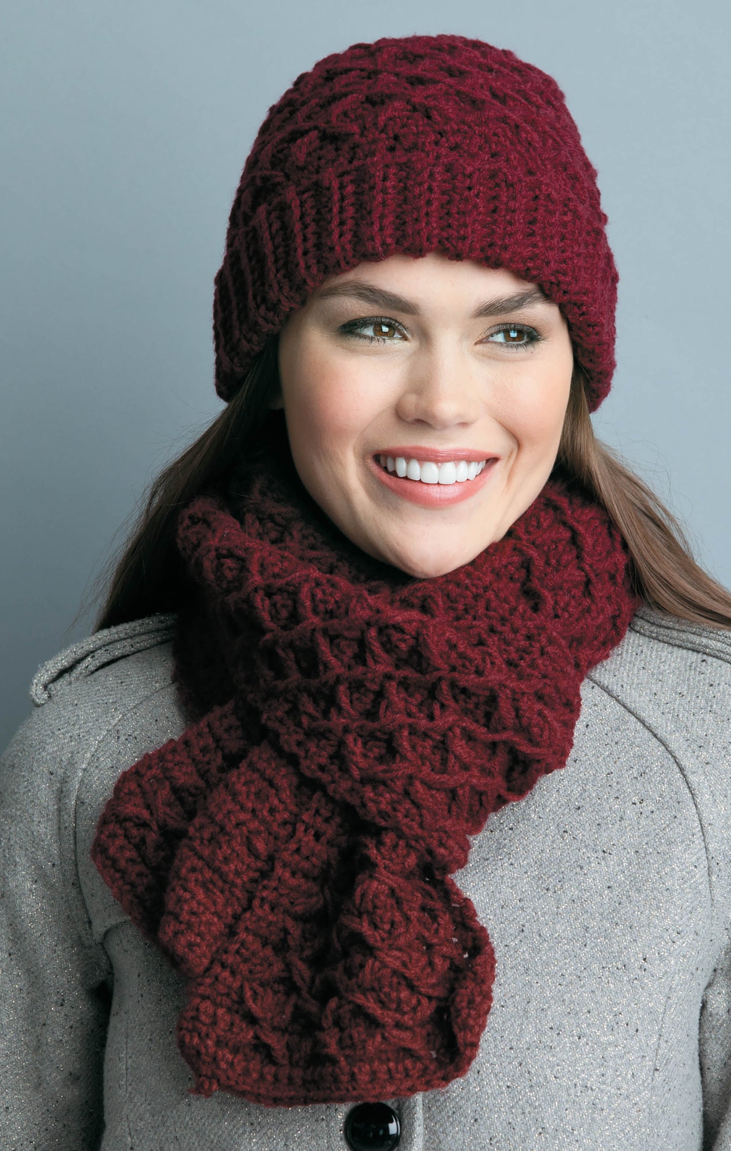 A woman models Leisure Arts’ Hats & Scarves for the Family, wearing a textured maroon knit set—ideal winter gifts—paired with a gray coat against a plain gray background.