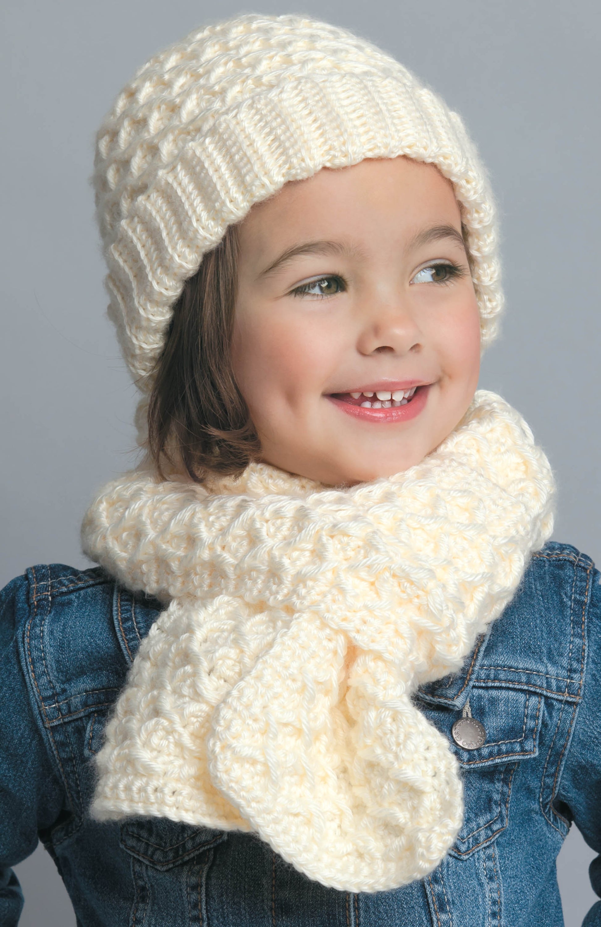 A child in a cream knit hat and scarf, smiling in a blue denim jacket against a gray background—showcasing Leisure Arts' "Hats & Scarves for the Family," perfect inspiration for winter gifts or cozy crochet patterns.