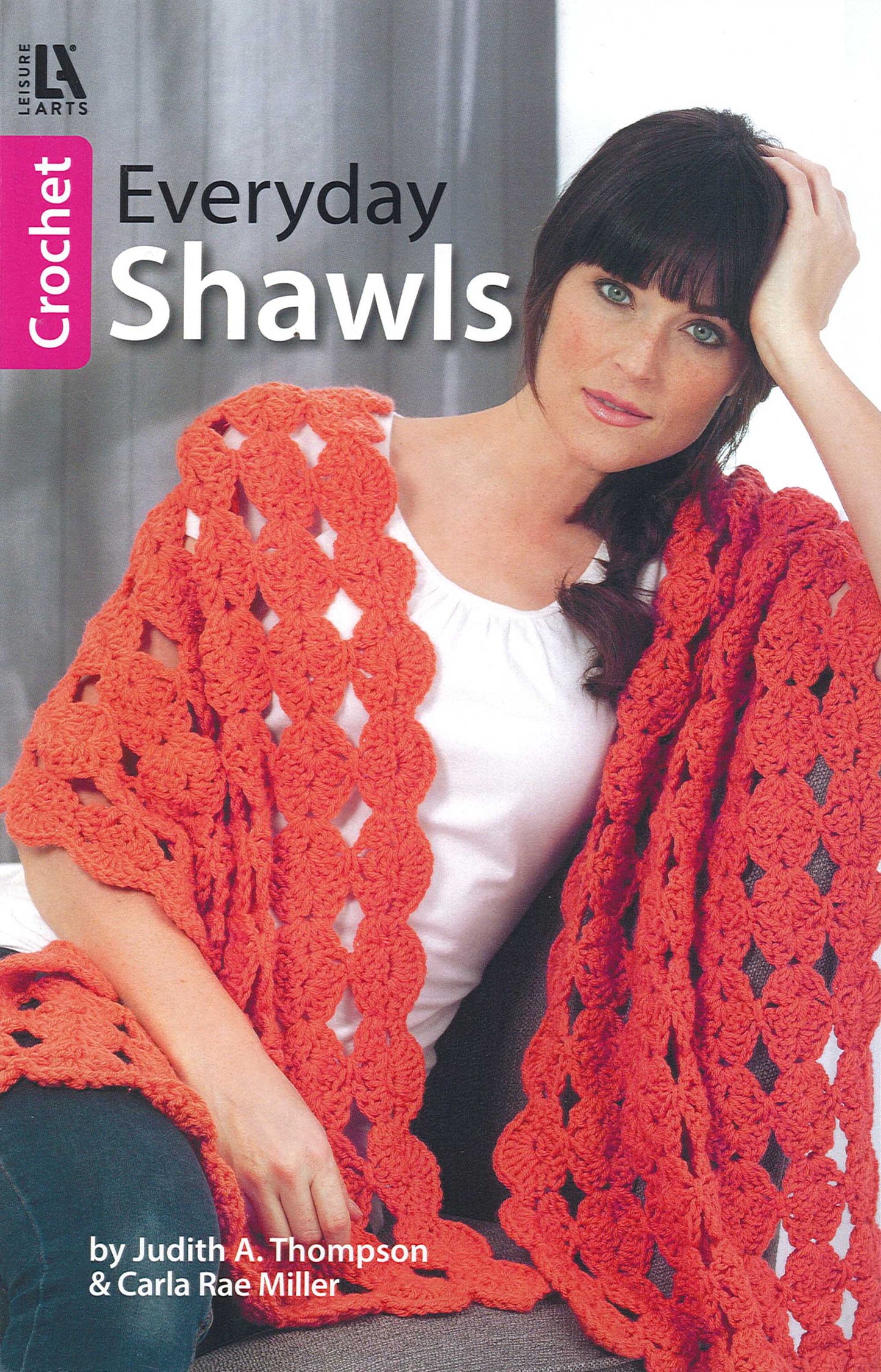A woman sits on a couch wearing a bright orange worsted weight yarn shawl. She holds her head in one hand and looks at the camera. The book “Everyday Shawls” by Leisure Arts is visible on the cover.