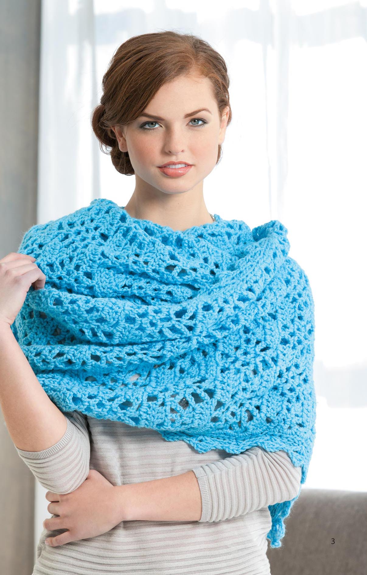 A woman with light skin and brown hair models the Leisure Arts Everyday Shawls—a large, bright blue worsted-weight crochet accessory—indoors before sheer white curtains; ideal for beginner crocheters.