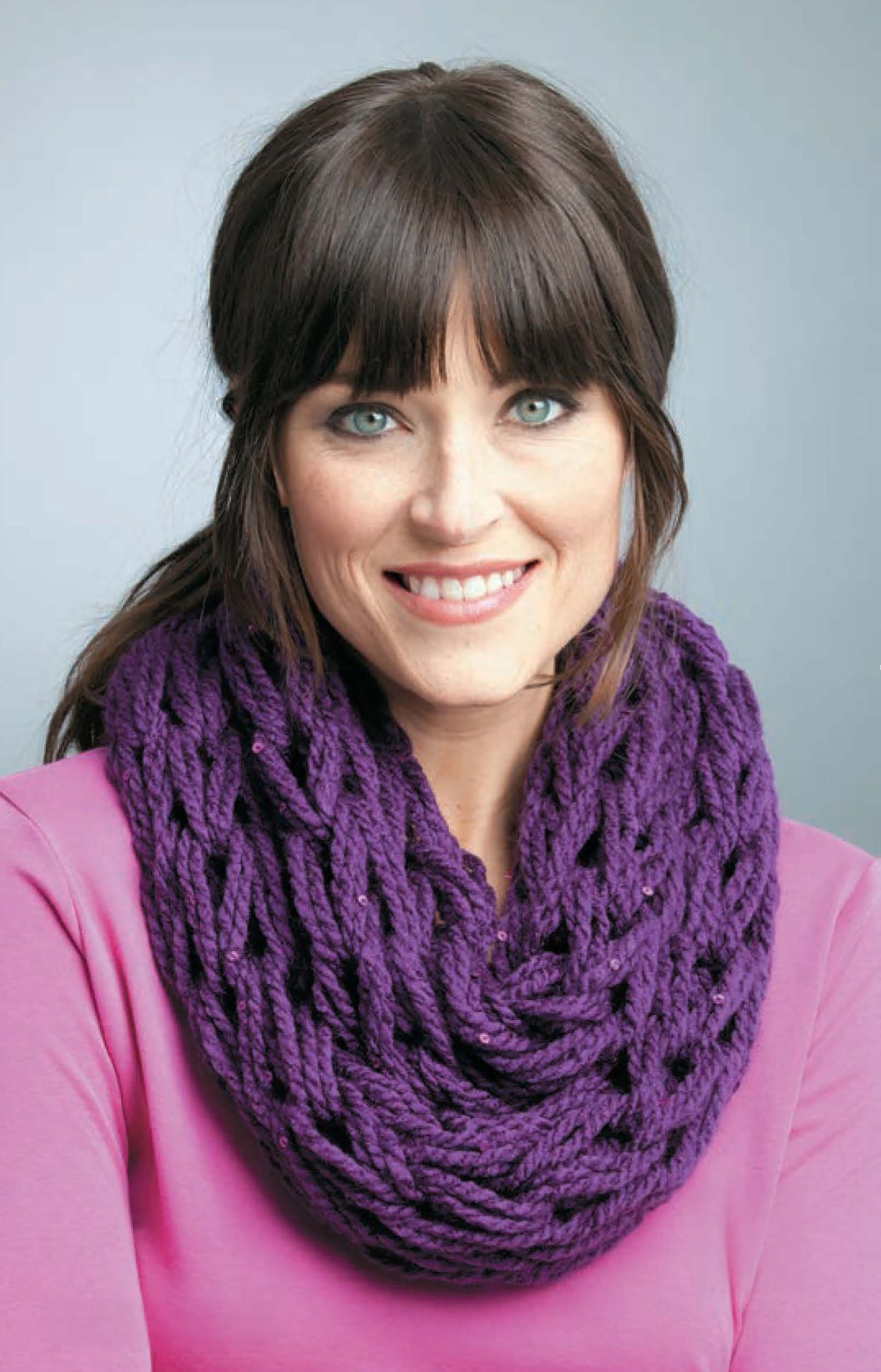 A smiling woman with straight brown hair and bangs models a chunky purple cowl made with "Learn to Arm Knit" by Leisure Arts. She wears a bright pink top against a light gray background.